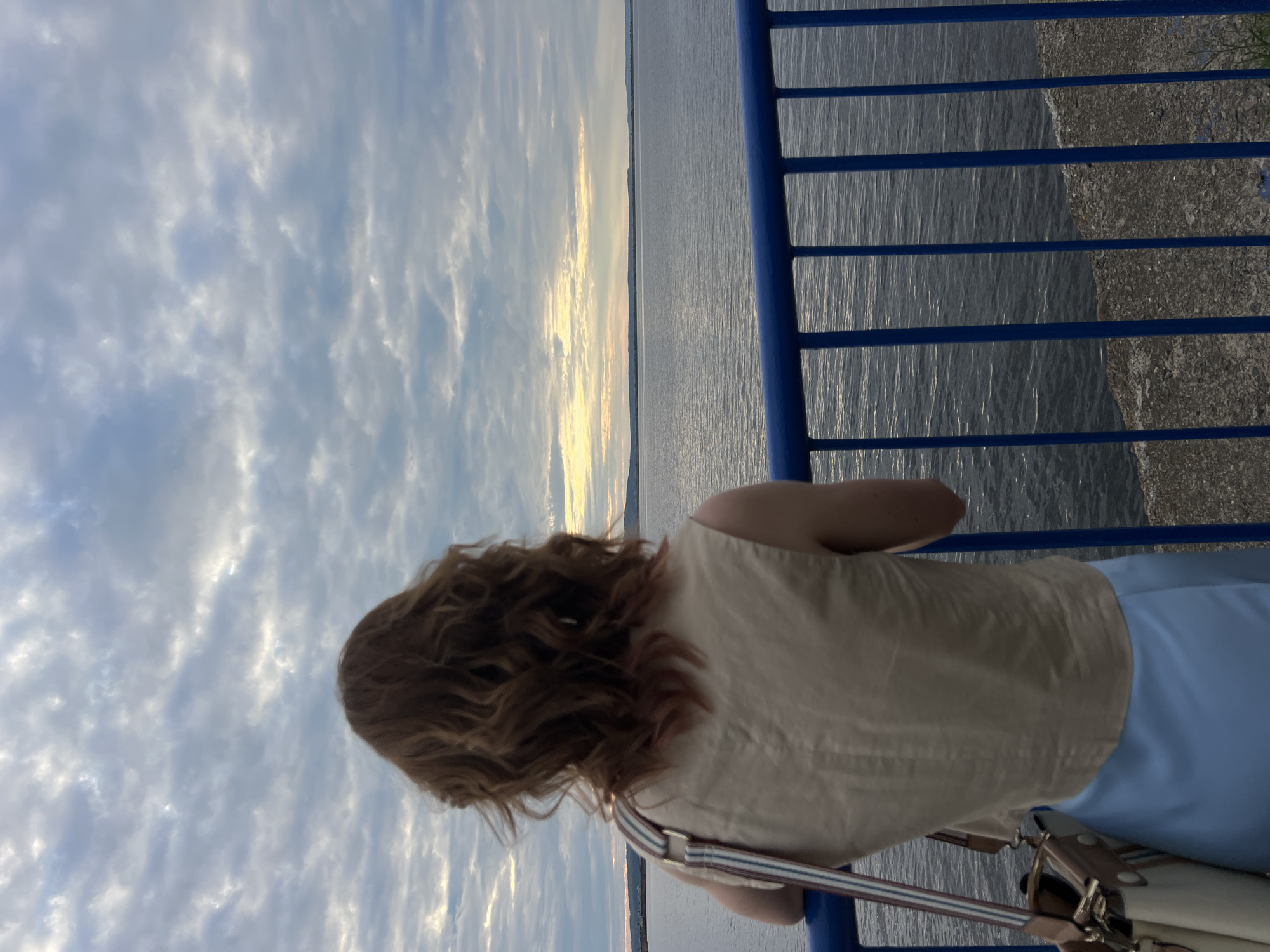 A woman with long, wavy auburn hair stands with her back to the camera, looking out at a body of water. She is wearing a light-colored, sleeveless top and light blue pants. A bag with striped straps is slung over her shoulder. She is leaning against a blue railing, which runs along the edge of a waterfront promenade. To the right of the railing, there is a stony ground. The water is calm, reflecting the sky and the soft light of a setting sun. The sky is filled with clouds, some of which are illuminated by the warm glow of the sunset. The overall scene evokes a sense of peace and contemplation. The time of day appears to be sunset. The location is likely a waterfront in Chaikovsky, Russia, given the context provided. There is no visible text in the image.