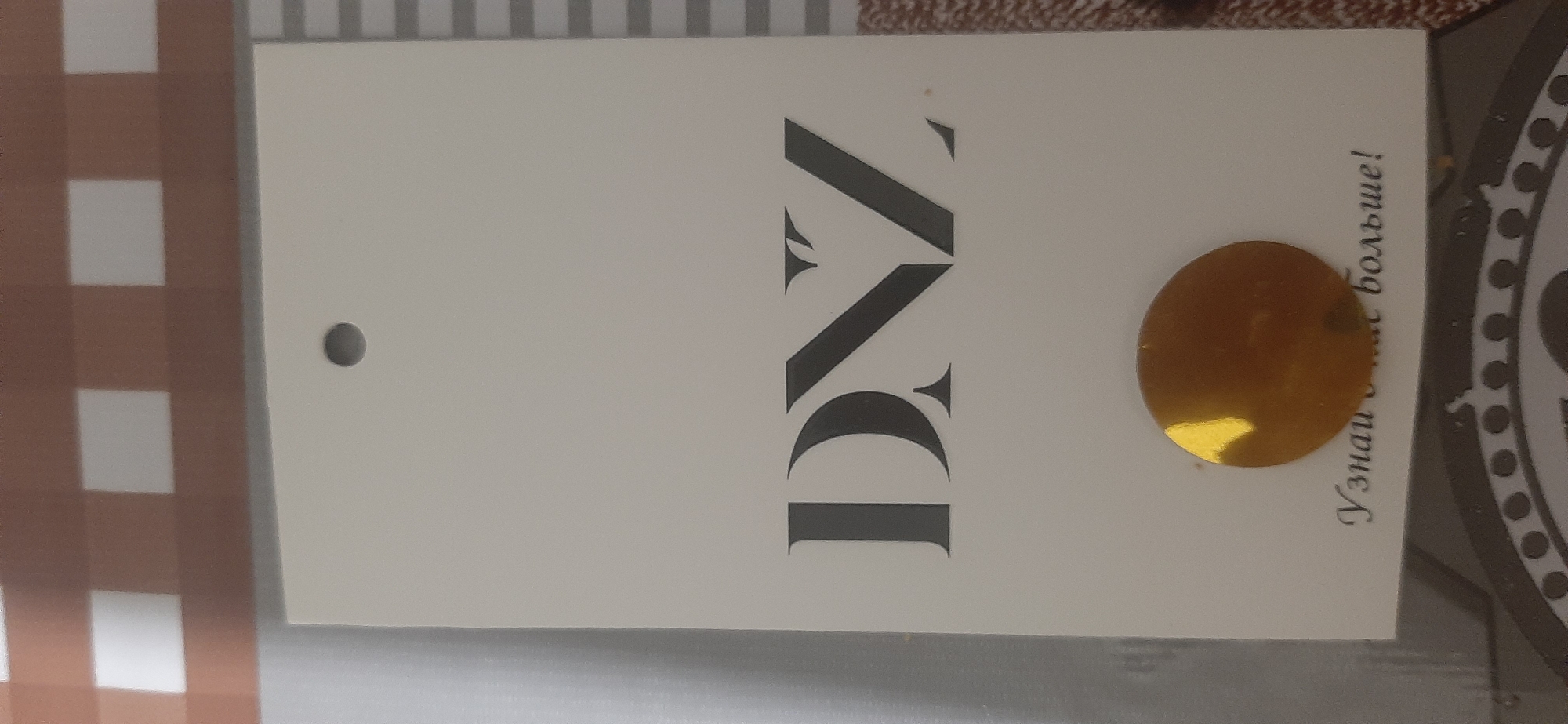 The image shows a white clothing tag with the logo "DNZ" printed in a dark, serif font. Below the logo, there is a gold circular sticker. Underneath the sticker, text in a cursive font reads "Узнай о нас больше!" which translates to "Learn more about us!". The tag is hanging, with a small hole at the top for attachment. The background is blurred but appears to be a tablecloth with a checkered pattern of brown and white squares. There are no people or discernible environmental features indicative of the city of Yessentukskaya, Russia.