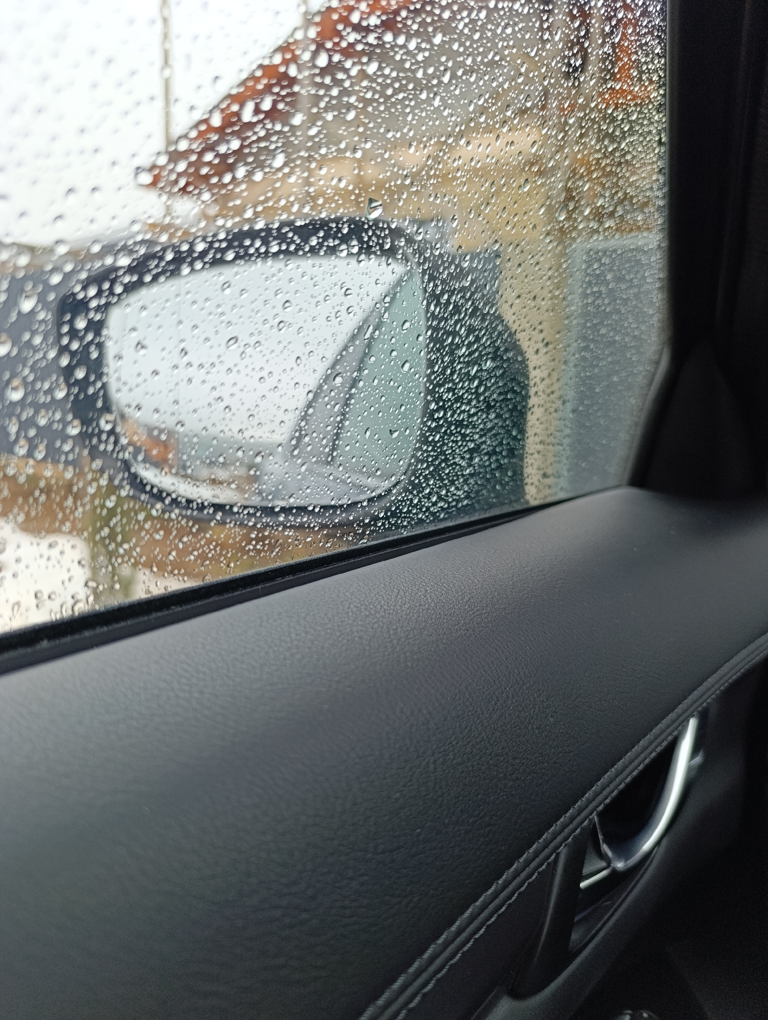 A close-up shot from inside a car shows a rain-covered window and side mirror. The exterior view is obscured by raindrops, but a building with a reddish-brown roof is partially visible. The interior includes a black leather door panel and a silver door handle. Location cannot be determined definitively as Chaikovsky, Russia from the given information.