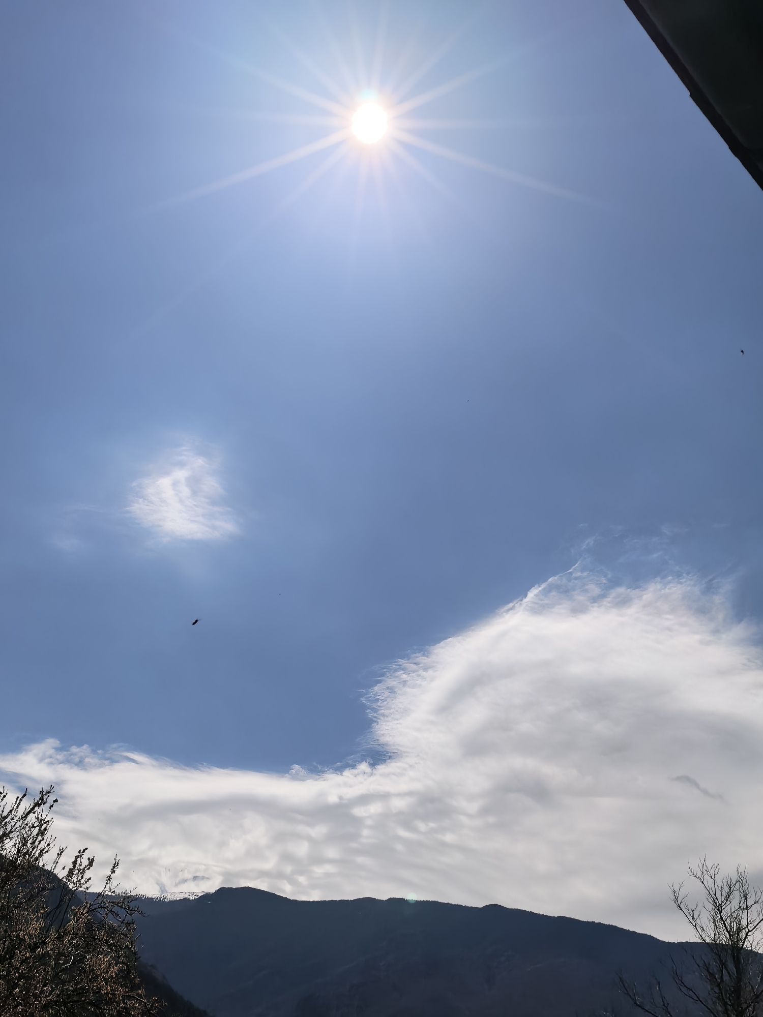 The image shows a daytime view of a sky with a bright sun at the top center, surrounded by sunbeams. The sky is predominantly blue with scattered clouds. In the lower portion of the image, there's a dark silhouette of mountains and vegetation on the left and right sides. One bird is flying in the sky. Location: Unknown, Unknown.