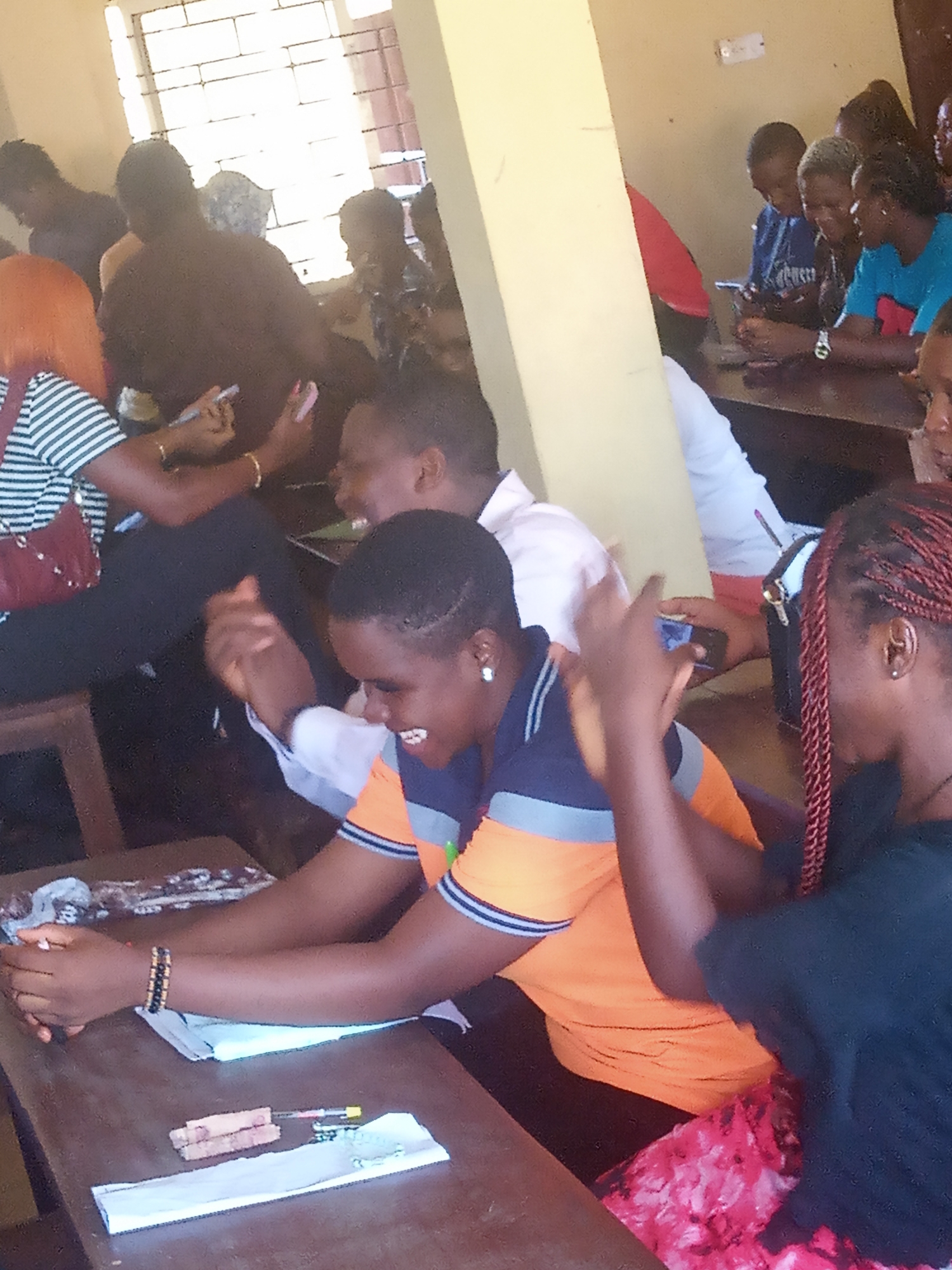 The image shows a classroom in Ukwu-Abwa, Nigeria. A group of young adults are seated at desks. Some are looking at their phones, while others are laughing and interacting with each other. One person in the foreground wears a blue and orange shirt and is laughing with their hands raised. A person with braided hair in a black shirt sits beside them. The desks appear wooden, with papers and other small items scattered on them. A window is visible in the background, providing natural light.