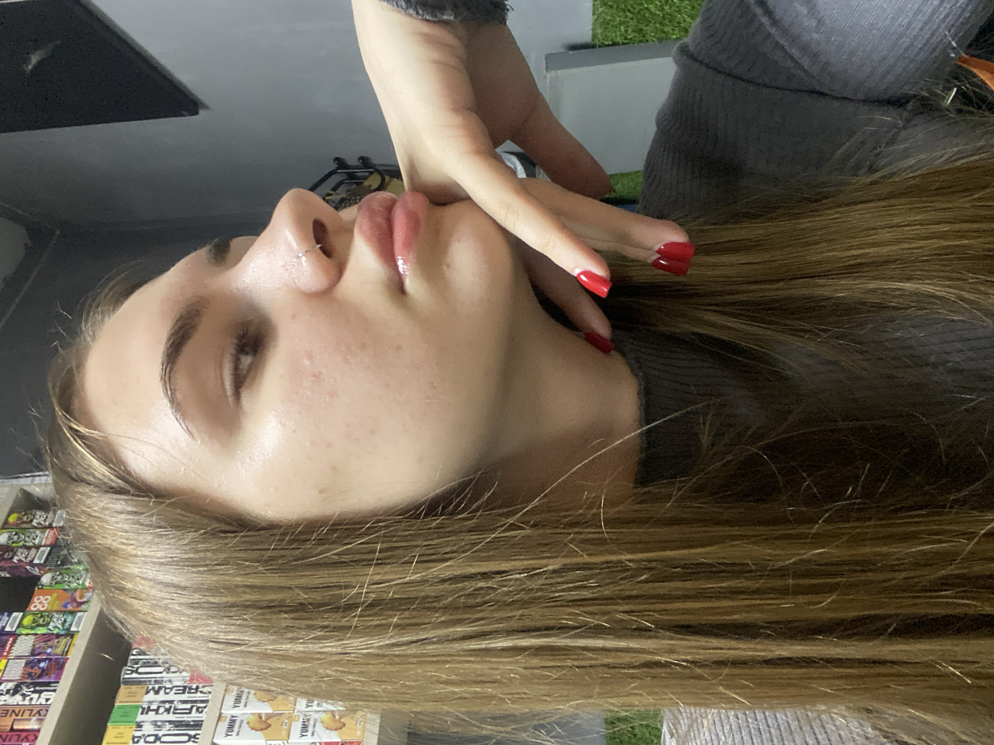 A young woman with long, blonde hair and a nose ring is pictured from the chest up, facing right. She is wearing a grey, ribbed long-sleeved shirt. Her left hand is raised, with fingers touching her chin and lower lip. Her fingernails are painted red. The background includes shelves stocked with colorful boxes, some appearing to be vape juice containers, with text visible such as "YUMMY" and "CREAM". There is also a hint of artificial green grass to the right. The overall setting appears to be an indoor retail or storage space. The city of Dyakovo, Russia is not depicted or referenced in this image.