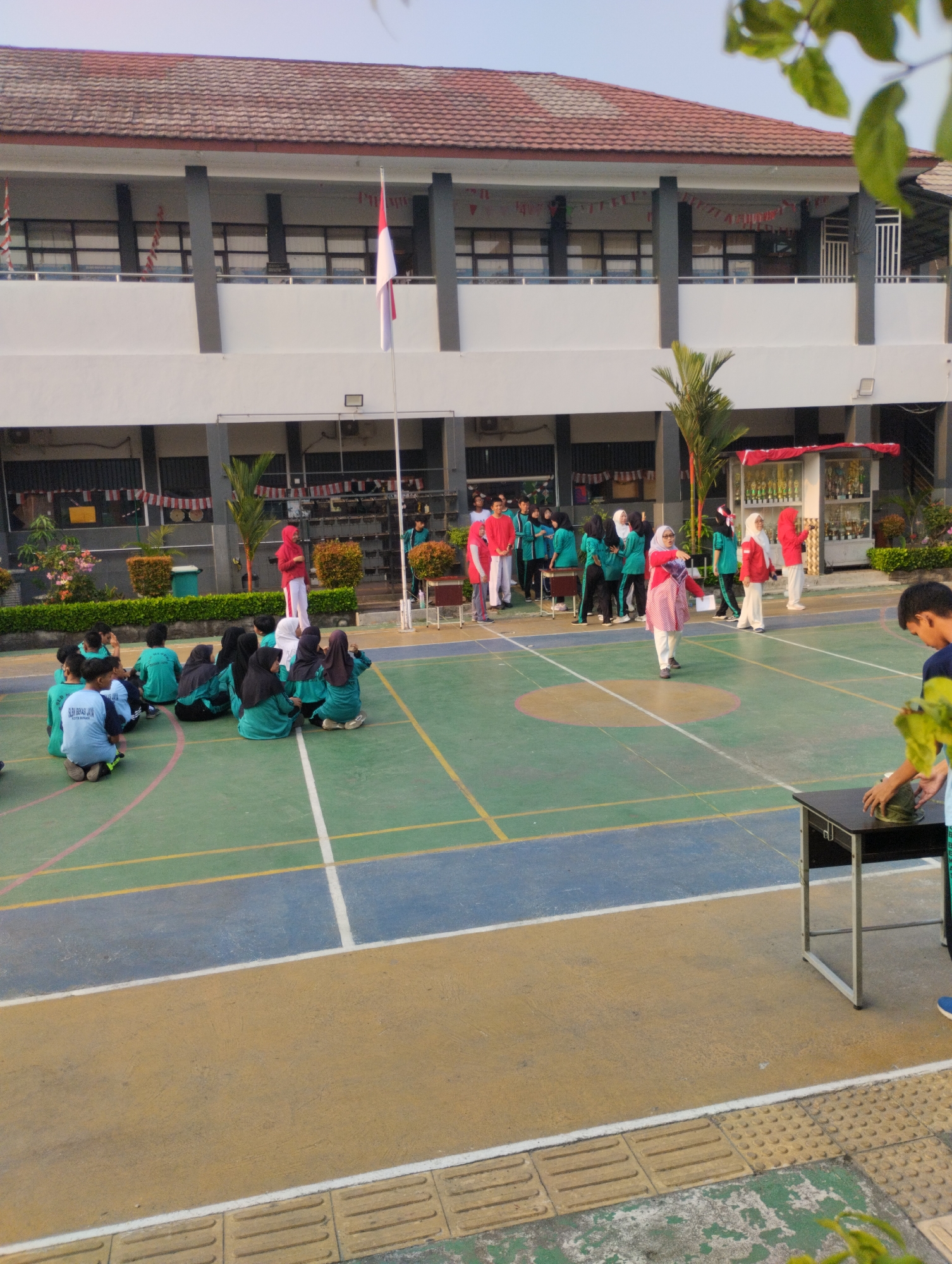 The image depicts an outdoor scene at a school in Bekasi, Indonesia, likely during a flag-raising ceremony or a school event. In the background, a two-story building with a red tiled roof and white and gray facade stands. A flag of Indonesia is prominently displayed on a flagpole in front of the building.
In the foreground, a group of students wearing green uniforms are sitting on a multicolored court, which appears to be a basketball or volleyball court, indicated by the painted lines. Some of these students are wearing hijabs. A few adults, some in red and white attire, are standing and interacting with students. One adult in a red and white patterned outfit is in the center, appearing to address or direct students. Another person, dressed in red, is standing to the right.
To the right of the court, a black table is positioned, and a young male student in a blue shirt is standing next to it, interacting with something on the table. The environment appears to be well-lit, suggesting it is daytime. The presence of potted plants and greenery around the building adds to the school campus setting. The court surface shows wear and tear, with visible scuff marks and faded paint. The overall atmosphere suggests an organized school activity is taking place.