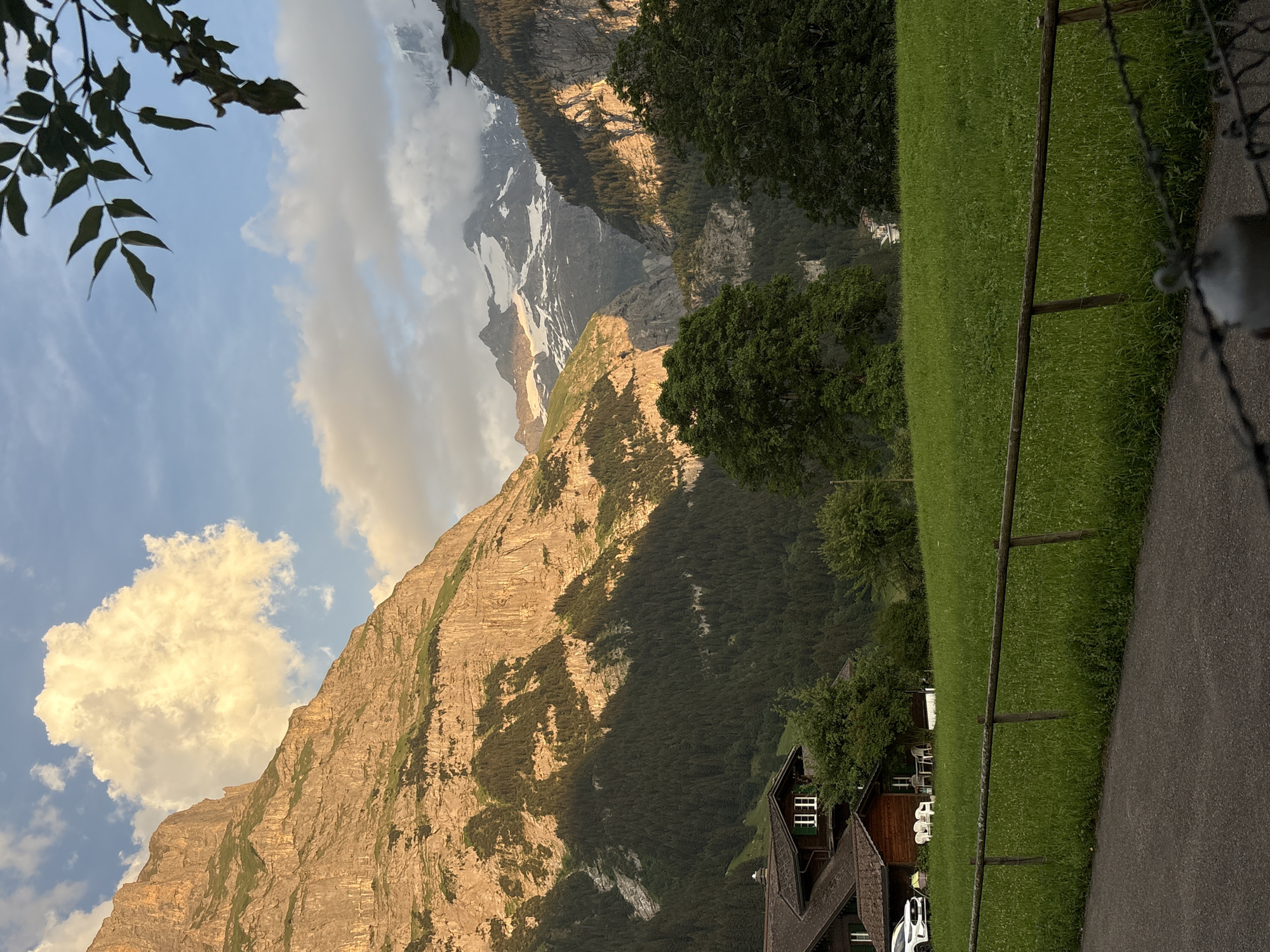This media file captures a scenic view of the Swiss Alps, likely in Grindelwald, Switzerland. The image is oriented vertically and appears to be taken during the late afternoon or early evening, as indicated by the warm, golden sunlight illuminating the mountainside. A large, rugged mountain face dominates the left side of the frame, with patches of green vegetation clinging to its slopes and areas of snow and rock visible higher up. The sky is a mix of blue and white clouds, with one prominent, brightly lit cumulus cloud.
In the foreground on the right, there's a neatly mown green lawn bordered by a rustic wooden fence. A paved path runs alongside the lawn, suggesting a park or residential area. A traditional Swiss chalet-style building with a dark wood exterior, white-trimmed windows, and a steep roof is partially visible at the bottom of the frame. Some outdoor chairs are arranged on the lawn near the building. Trees, both deciduous and coniferous, are scattered throughout the scene, adding to the natural beauty. The overall impression is one of tranquility and the majestic grandeur of the alpine landscape. No people or specific events are discernible, focusing entirely on the natural and architectural setting.