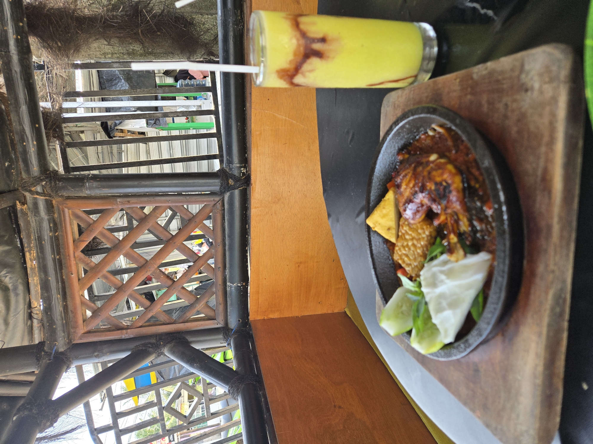 A close-up, eye-level shot focuses on a meal served on a wooden plank on a black table, likely at an outdoor eatery in Parit Padang, Indonesia. To the left is a sizzling platter containing what appears to be grilled chicken or fish in a dark, rich sauce, garnished with cucumber slices, shredded cabbage, and a piece of fried tempeh. To the right, a tall glass of yellow smoothie, drizzled with chocolate syrup, stands with a white straw. In the background, a rustic bamboo structure forms a ceiling and railing, with glimpses of other patrons and structures through the openings. The overall setting suggests a casual dining experience, possibly under a sheltered outdoor area.