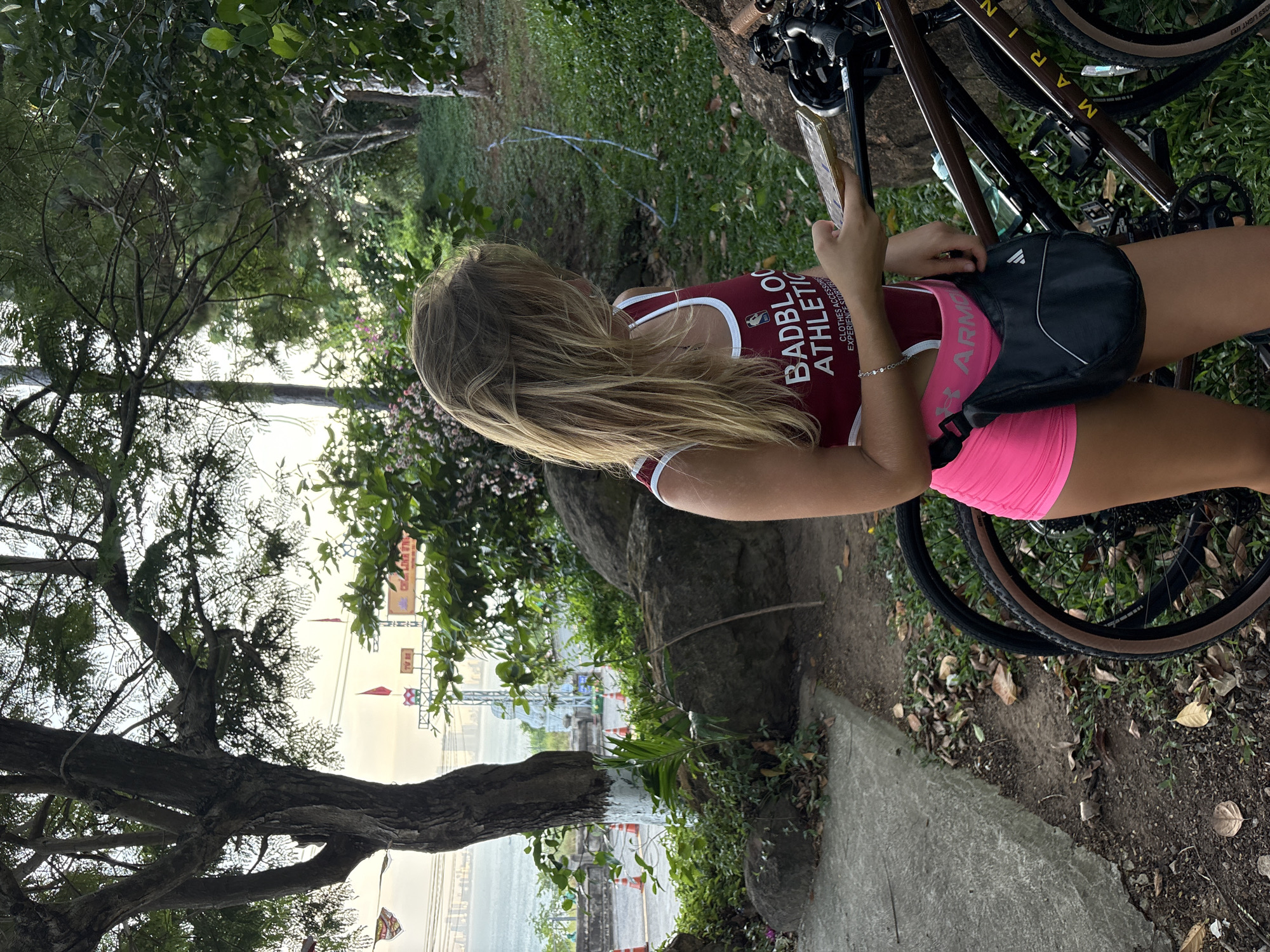 A woman is standing next to a bicycle on a paved path with vegetation in Đà Nẵng, Vietnam. The woman has blonde hair and is wearing a maroon and white tank top with the words "Badblood Athletic" printed on it and pink shorts. She is holding a phone in her hand. The bicycle is a dark brown color with a brown seat. The environment includes trees and vegetation. In the distance, there's a body of water and buildings, including decorations like flags. A tree trunk is painted white.