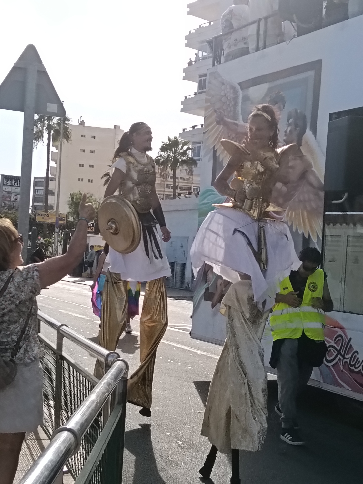The image captures a vibrant street scene in San Bartolomé de Tirajana, Spain, possibly during a parade or festival. Two individuals on stilts are prominently featured. The first, a male, is dressed in a golden breastplate, white tunic, golden leggings, and carries a golden shield. The second, likely a female, is adorned with a golden breastplate, wings, and a white skirt, also on stilts covered in a white and silver cloth. Behind them is a truck with a mural of angels.
A woman on the left side of the frame is gesturing with her hand raised, possibly interacting with the stilt walkers. A man in a yellow safety vest stands near the truck. The background includes buildings, palm trees, and street signs, providing context for the urban location. The presence of a "Habitat" sign and "MOTO" sign are visible. The weather appears sunny and bright.