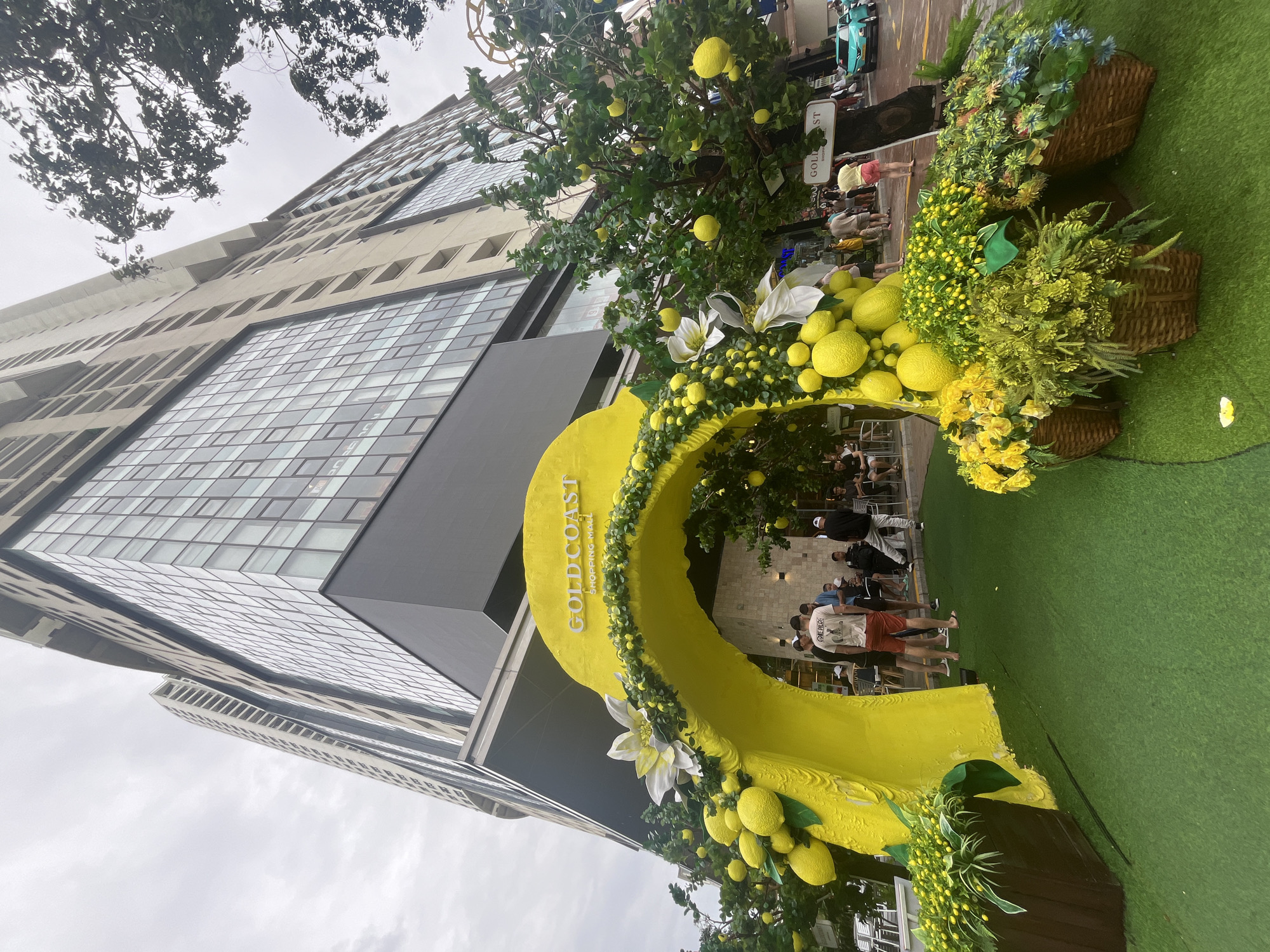 The image depicts an outdoor scene in the city of Unknown, Unknown. A large, yellow, lemon-shaped archway, decorated with artificial lemons and flowers, stands on a patch of artificial green turf. The archway is labelled "GOLD COAST SHOPPING MALL". People are walking through and standing behind the archway. Baskets of yellow and blue flowers are visible to the right of the archway. In the background, a tall, modern building with a glass facade is partially visible, with overcast skies above. A tree with fake lemons hangs on the front.