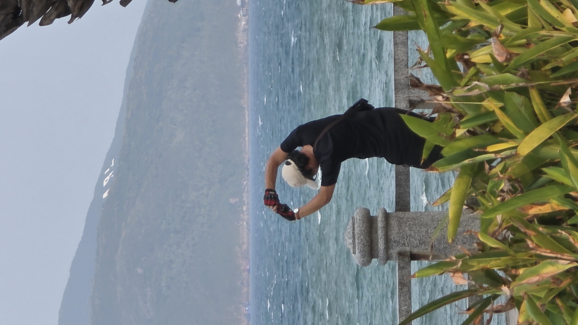 A man in a black shirt, black shorts, a tan cap, and red and black gloves is bending over a stone railing near a body of water. He appears to be stretching or exercising. In the background, a mountain with what appears to be white writing on its side is visible. Green plants surround the railing. The body of water is likely the sea. The location is the city of Nha Trang, Vietnam.
