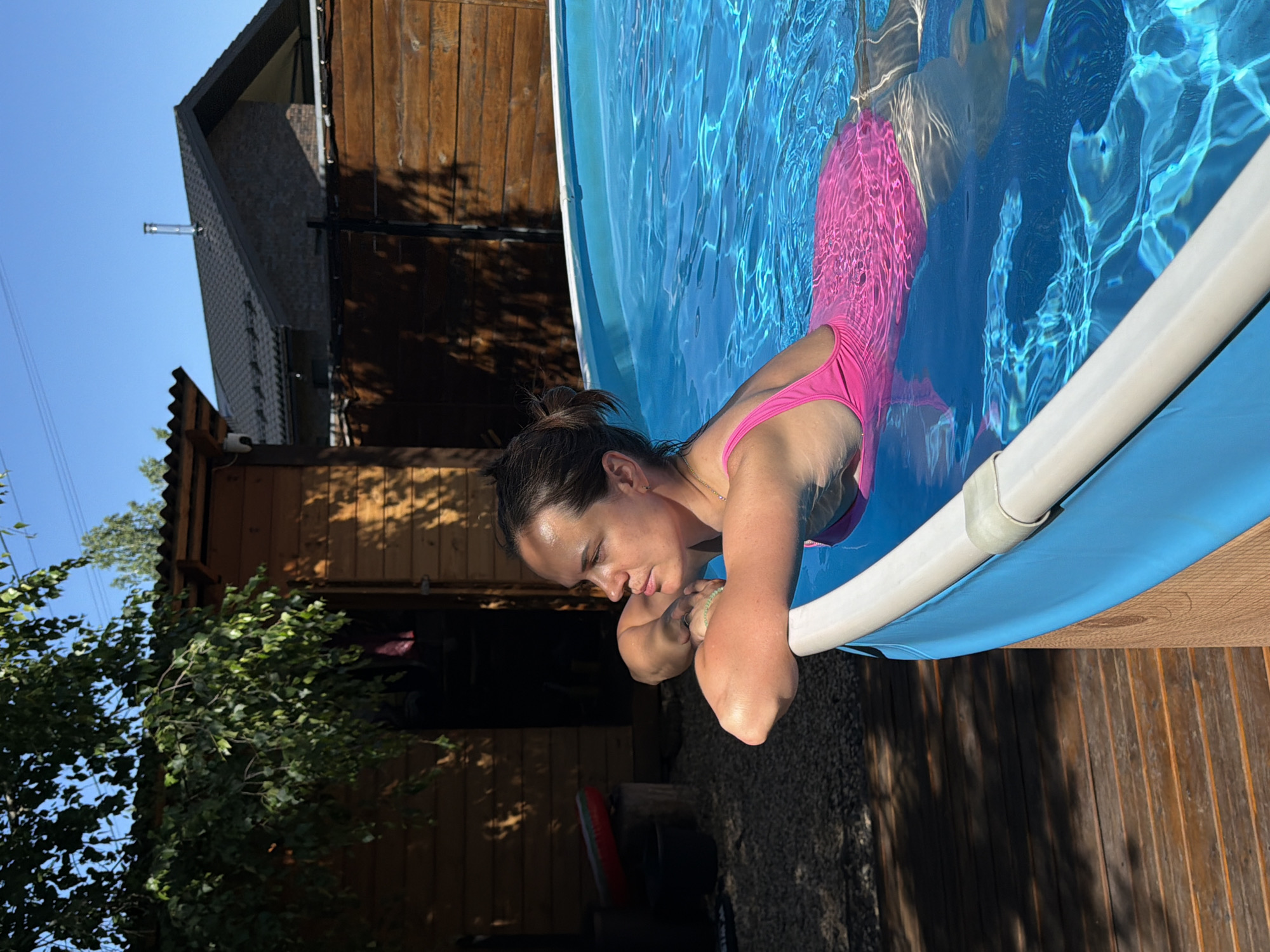This image captures a sunlit residential backyard scene in Chertovitsy, Russia, featuring a woman in an above-ground swimming pool. The central figure, a woman with dark hair pulled back, is seen in profile from the chest up, leaning with her forearms on the white rim of a blue-lined pool. She wears a vibrant pink one-piece swimsuit, and her skin appears tanned under the bright sunlight. The pool water is clear, blue, and shows ripples reflecting the light.
To the left and behind the woman, a wooden shed or small building with a dark, corrugated roof is visible. Green leafy tree branches partially obscure the shed's roof and cast dappled shadows across its wooden facade and the ground below. A dark opening, possibly an ajar door, is seen on the shed. Near the shed, a red and black inflatable object rests on a wooden stump, and a dark bucket is on the ground, which transitions from gravel to wooden decking in the foreground.
In the mid-ground and right, a horizontal wooden fence separates the pool area from a residential house. This house features a dark, possibly tiled, sloped roof with a visible metal chimney or vent pipe. Its walls are constructed of dark brick or stone. The sky above is clear and blue, with faint utility lines visible. The scene is illuminated by strong, direct sunlight, creating distinct shadows across the landscape.
