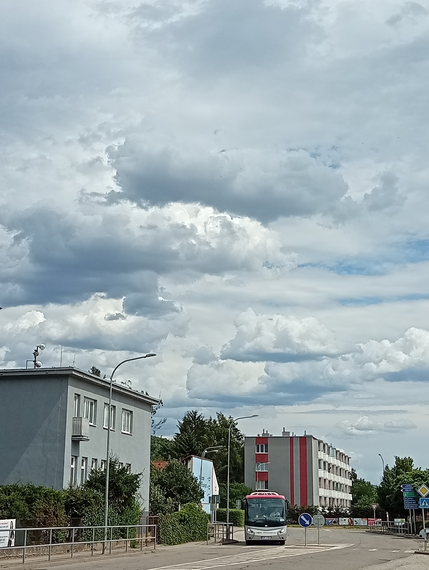In Tišnov, Czechia, on a cloudy day, a silver and pink bus drives forward on a paved road, crossing a pedestrian crossing. The bus has its headlights on and appears to be occupied by passengers. To the left of the road, a gray building with white-framed windows and a small balcony is visible, partially obscured by lush green bushes and trees. A street lamp stands in front of this building. Further down the road, to the right, a multi-story building with a gray facade and red vertical stripes is prominent. Several street signs are also visible on the right side of the road, indicating directions and traffic rules. The sky is filled with dramatic, billowing clouds, suggesting potential for rain.