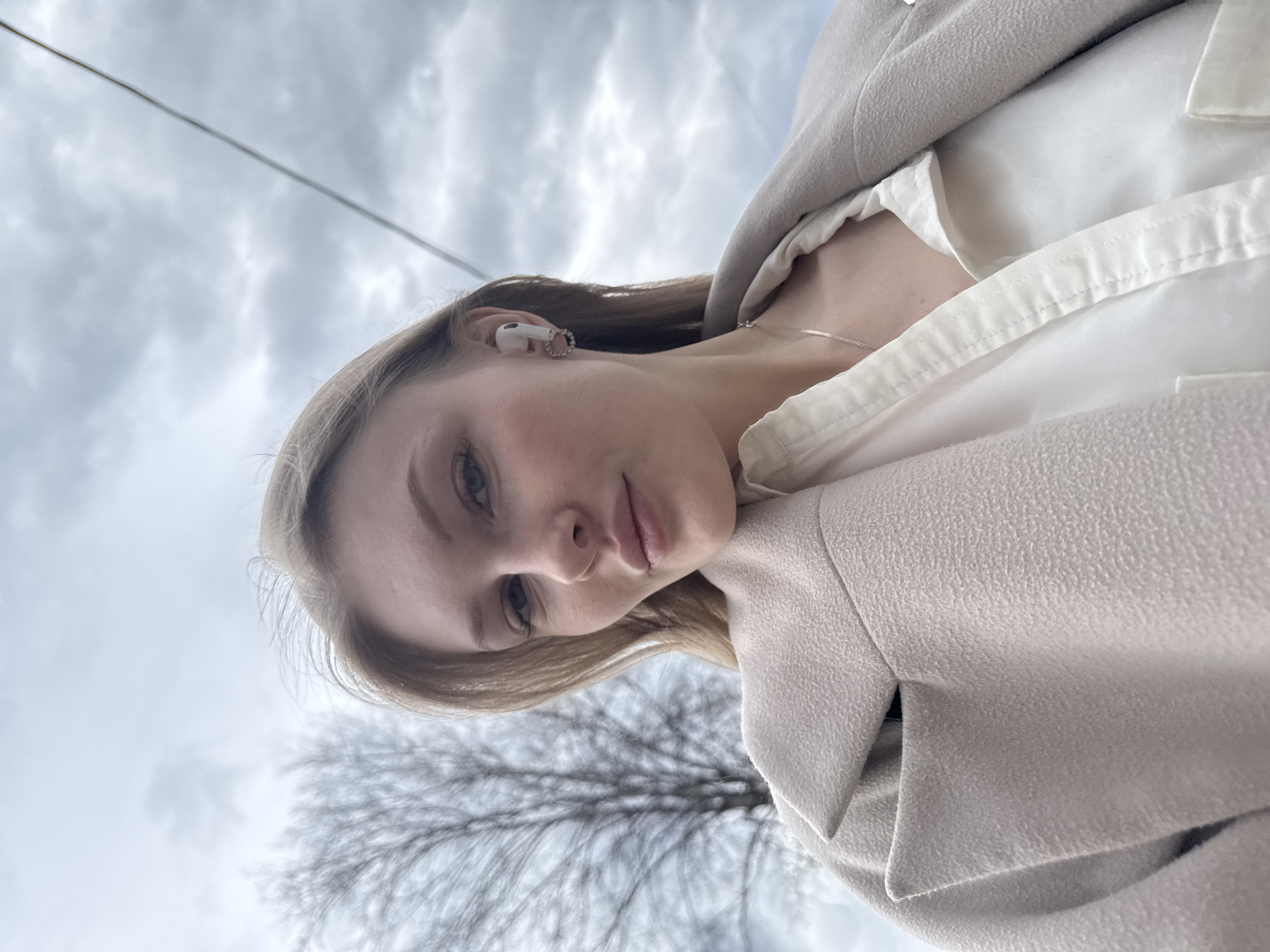 A low-angle shot shows a fair-skinned young woman with blonde hair, fair skin, and an AirPod in her left ear. She wears a light-colored collared shirt and a beige coat. A thin silver necklace is visible around her neck. The background consists of a cloudy sky with a bare tree visible. A power line stretches across the upper left corner of the image. The image appears to be taken from a ground-level perspective, facing upwards. The location is inferred to be in Perm, Russia.
