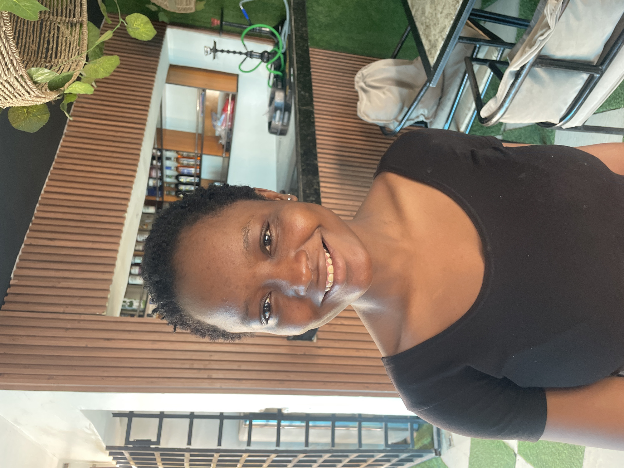 In Jos, Nigeria, a young woman with dark skin and short, curly hair is pictured smiling directly at the camera. She is wearing a black t-shirt. The background features a modern establishment with wooden slatted walls, a bar area with bottles and shelves, and artificial grass. To the left, a window with a metal grate is visible, and to the right, a hookah pipe and tables with chairs can be seen.