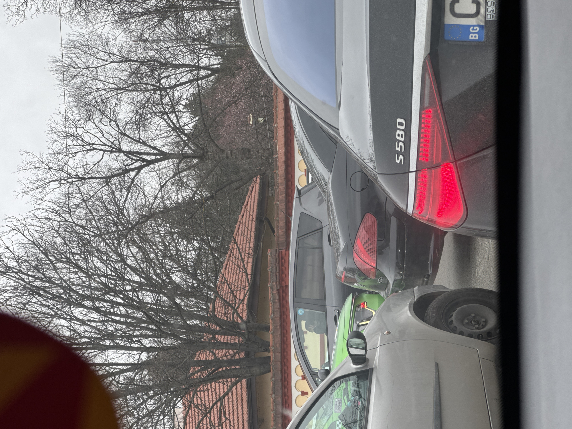 The image shows a close-up of several cars in traffic on a street. The closest car is a grey sedan, followed by a green car, and then a black Mercedes S580, indicated by the badging on its trunk. The Mercedes has a Bulgarian license plate. The location appears to be an urban environment, likely in Sofia, Bulgaria, due to the Bulgarian license plate. Bare trees are visible in the background, along with a building with a red tiled roof. The overall weather condition appears overcast. A portion of what appears to be a red and yellow sign is visible in the lower left corner of the frame.
