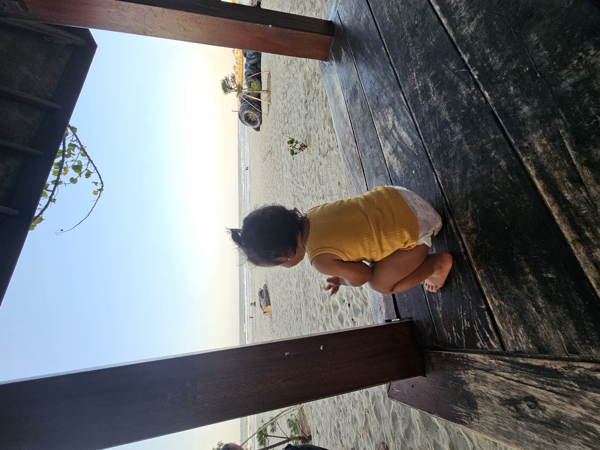 A toddler sits facing away from the camera, on a wooden structure on a sandy beach. The toddler is wearing a yellow tank top and a diaper. The beach is wide, with a gentle surf rolling in from the ocean. In the background, there are some logs, a small tree, and a pile of tires. The sky is clear and blue, with a hint of sunset colors on the horizon. The scene appears to be at the Dalegan beach in Gresik, Indonesia, a popular tourist spot known for its laid-back atmosphere and natural beauty.