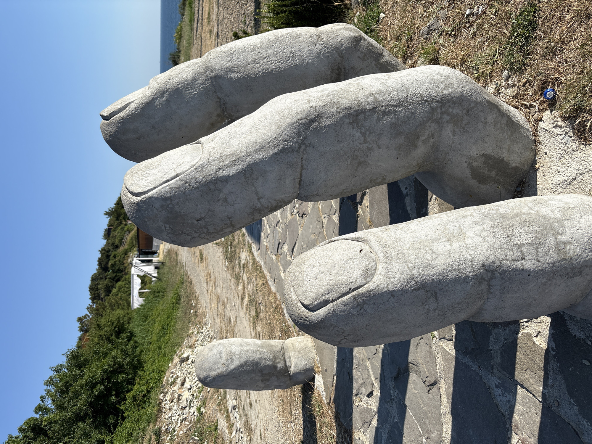 This media file is an outdoor photograph featuring a large, concrete sculpture of a hand with fingers partially visible. The sculpture appears to be made of rough, textured concrete and is placed on a surface paved with irregular stones.
The scene is set outdoors, under a clear blue sky, suggesting a bright, sunny day. The background shows a grassy area with dry vegetation, some trees and bushes, and in the distance, a body of water, likely the sea. There is also a glimpse of a building structure with white fabric, possibly a tent or canopy, nestled among the greenery on a hillside. The lighting indicates it is daytime, with distinct shadows cast by the sculpture.
The primary subject is the stone hand sculpture. The focus is on the texture and form of the concrete fingers, with visible fingernails. The perspective is close-up, emphasizing the scale and detail of the artwork. A small, blue and white object, resembling a bottle cap or a small toy, is visible on the ground near the sculpture, providing a sense of scale and hinting at human presence.
No visible text is present in the image. The overall impression is one of a sculpture placed in a natural, outdoor environment, possibly a park or a scenic overlook, in Kabardinka, Russia. The weather appears to be warm and sunny.