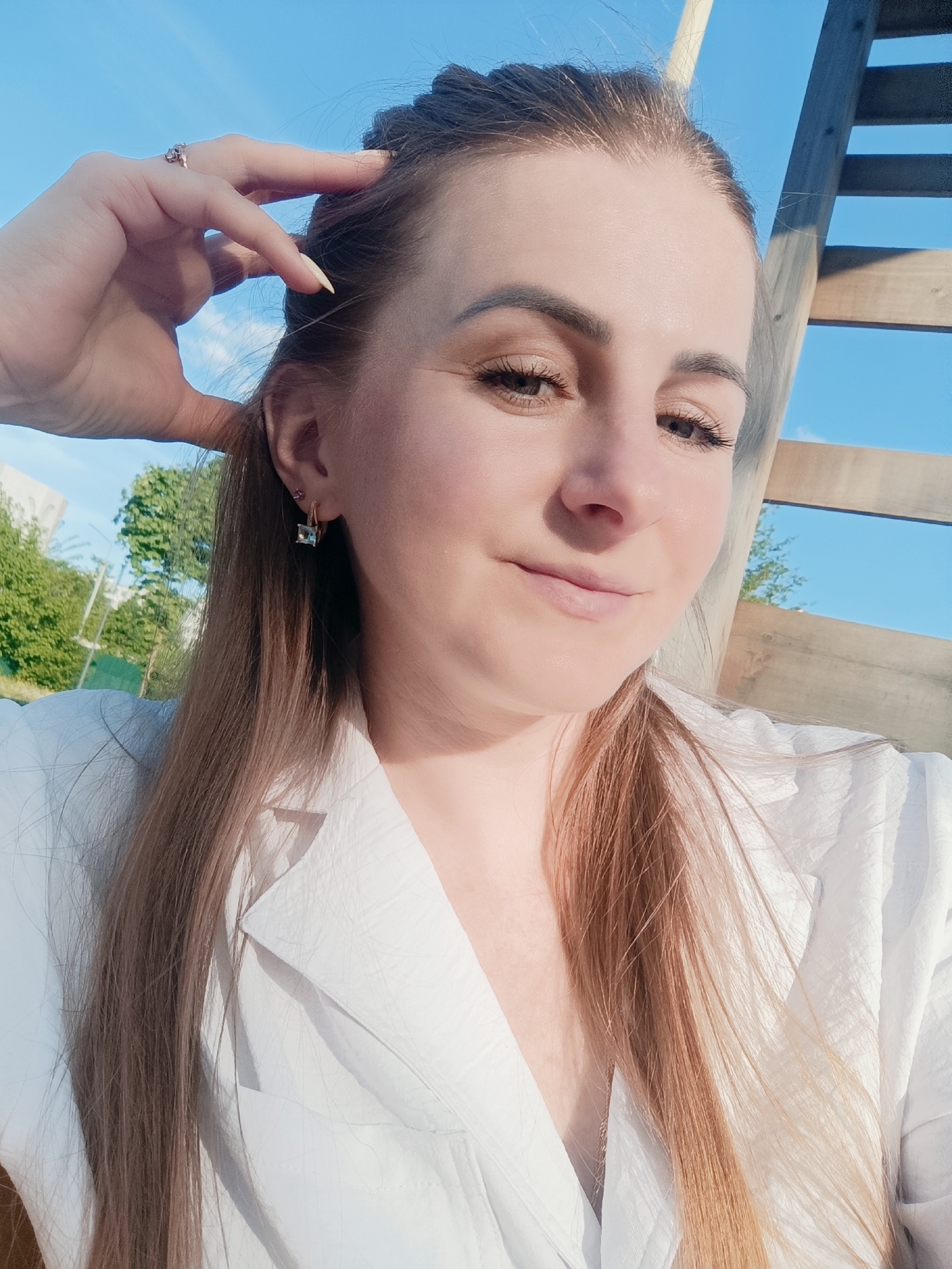 A woman with long, light brown hair poses for a selfie. She is wearing a white blazer-style shirt. Her left hand is raised, with her fingers touching her hair near her temple. She has fair skin and is wearing subtle makeup. A ring is visible on her finger and earrings on her ears. The background features a blue sky and a wooden structure, possibly a fence or a part of a playground. Trees and a building are visible in the distance. The location appears to be outdoors, possibly in a park or residential area.