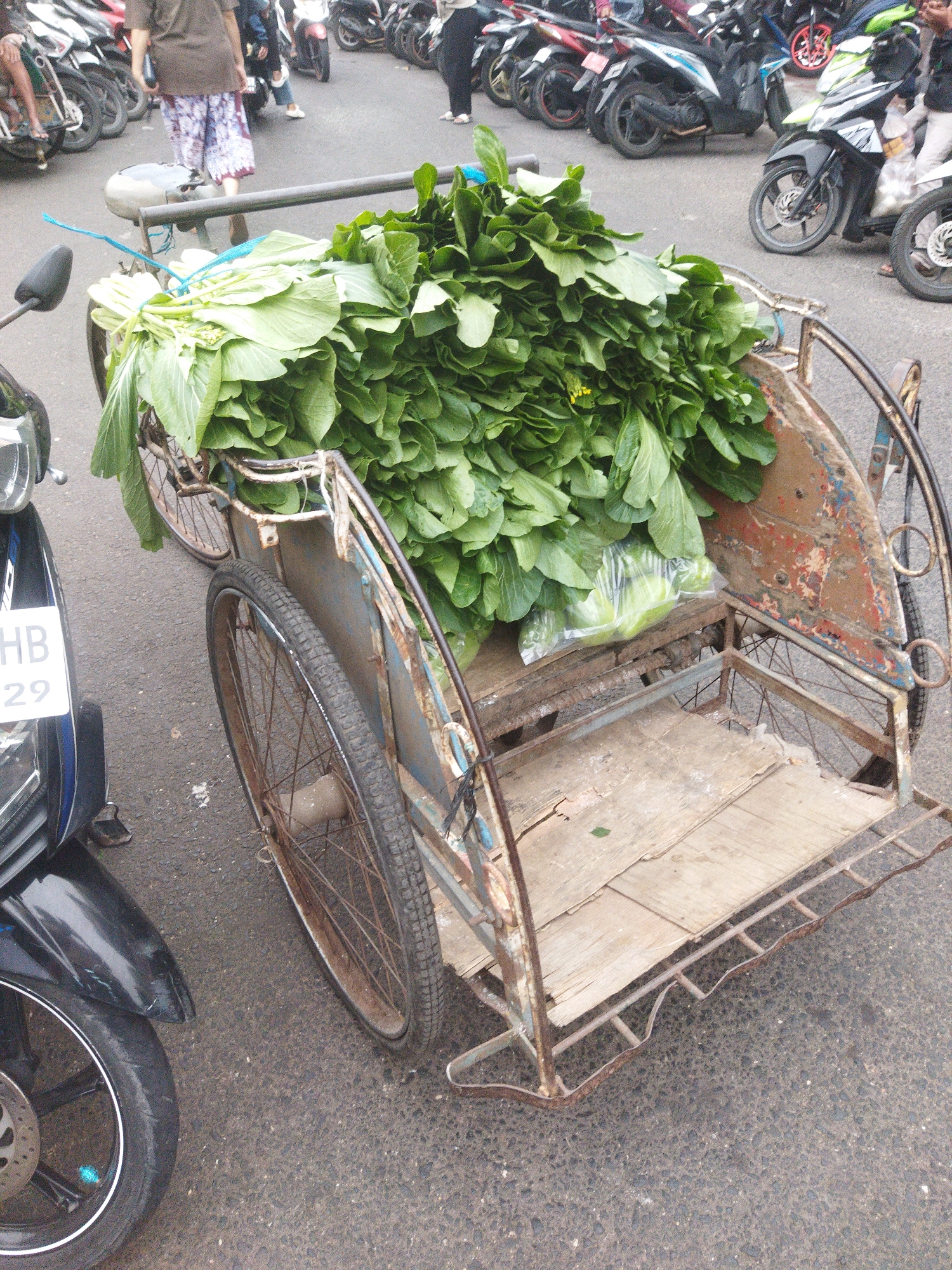 In Banjar, Indonesia, a rusty, three-wheeled cart laden with large bundles of leafy green vegetables, likely bok choy, is parked on an asphalt surface. The cart has a weathered, blue-painted metal frame with a wooden platform in its bed. In the foreground to the left, the front of a black motorcycle is visible, with its license plate displaying "HB 29". Behind the cart, several motorcycles are parked, with some people standing near them. One person, wearing a brown shirt and patterned pants, is partially visible walking away from the cart. The overall environment appears to be an outdoor market or street.