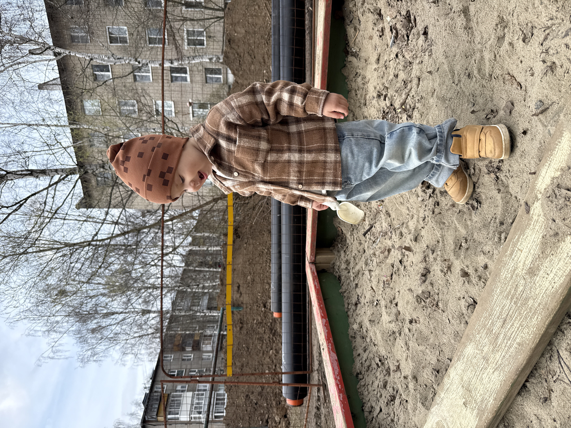 An outdoor scene depicts a young child playing in a sandy or dirt area, with urban buildings and bare trees in the background. The child, likely a toddler, is dressed in a brown plaid jacket, light blue jeans, and light brown boots, topped with a brown knit hat featuring a grid pattern. They are standing upright, holding a small white shovel in their right hand, seemingly engaged in play.
The immediate foreground consists of a mixture of sand and dirt, with a pale, weathered wooden plank visible on the right. A green-painted border and a red-painted rail separate the sandy area from a trench-like section containing two large black pipes. A metal fence, part brown and part yellow, runs along the mid-ground.
In the background, behind the child and the fencing, several multi-story apartment buildings with many windows are visible through the branches of bare trees. The lack of leaves on the trees suggests a season of late autumn, winter, or early spring. The sky is overcast, providing soft, diffused daylight, indicating it is daytime. The setting appears to be a residential or public open space in Rusanovo, Russia. There is no visible text in the image.