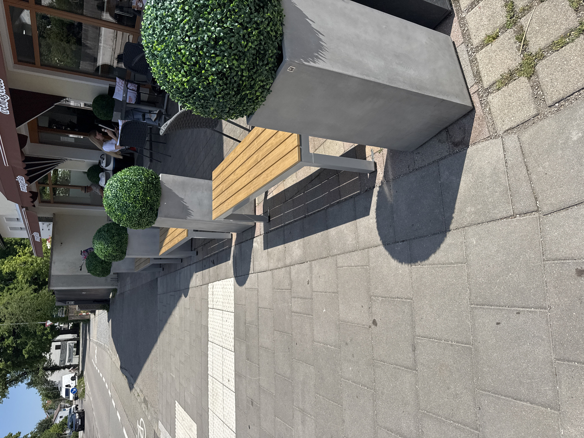 This is a vertical, eye-level shot taken outdoors on a sunny day, likely in the afternoon given the long shadows. The scene appears to be the outdoor seating area of a cafe or restaurant in Höhenkirchen-Siegertsbrunn, Germany, adjacent to a street.
The foreground is dominated by a paved sidewalk composed of rectangular gray stones. There are two distinct sections of paving: one with a rougher texture and embedded grass, and another with smoother, more regular patterns. A crosswalk with tactile paving is visible.
In the mid-ground, there are several modern benches with wooden slats and grey concrete planter boxes filled with spherical, artificial green foliage. One planter box is particularly large and prominent, casting a distinct circular shadow on the sidewalk. Behind the benches, the facade of a building with large windows and an awning can be seen. A person is seated at a table inside, looking at their phone.
The background shows a street with parked cars and trees lining the sidewalk. A white van is visible further down the street, along with a blue traffic sign and a bicycle symbol on the road.
The overall impression is one of a quiet, pleasant outdoor space, likely a popular spot for patrons of the establishment. The sun is bright, suggesting good weather. No discernible text is visible beyond what appears to be a small logo on the large planter box.