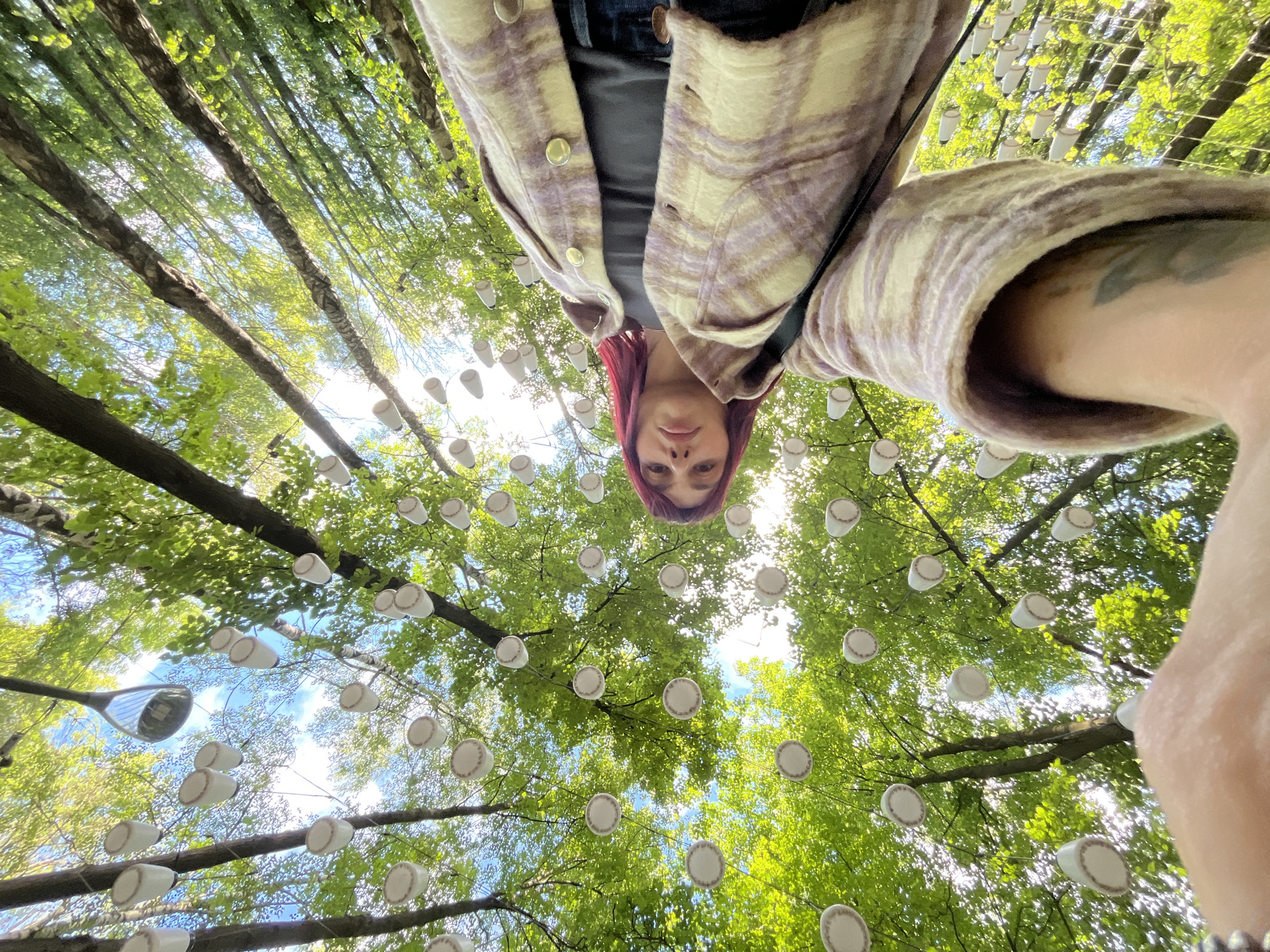 The image shows a low-angle, wide shot of a person taking a selfie outdoors. The person has reddish-pink hair and is wearing a plaid jacket over a dark top and jeans. They are looking upwards, with their face partially visible. The background is dominated by trees with bright green foliage and a blue sky with some clouds. Many white cups with brown decorative rims are hanging from the tree branches, creating an artistic installation. The scene appears to be set in a park or wooded area. The specific location in Nizhny Novgorod, Russia, is not identifiable from the visual information alone.