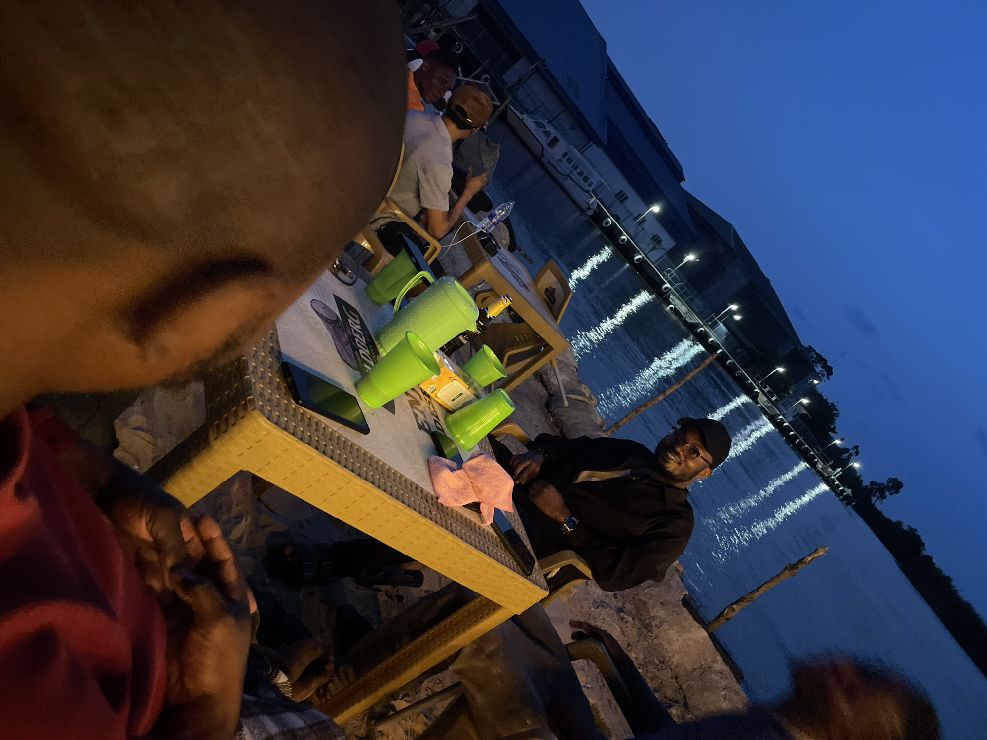 An evening social gathering is captured from an overhead, tilted perspective, showing a group of people seated by a body of water at dusk or night. The camera's subject, seen in the blurry foreground on the left, wears a red shirt, with hands visible in the lower left.
In the middle ground, several people are seated around light-colored, possibly rattan-style tables. On the left side of the main table, two individuals are partially visible, one in a light grey shirt and cap facing away from the camera. The central figure, seated to the right of the table, is a man with a black top, black baseball cap, and glasses, smiling directly at the camera.
The main table in the foreground-middle holds several bright green plastic pitchers and cups, a pink cloth, and a smartphone. A white and blue banner or mat on this table displays partial text, "LDERBERG," likely part of a brand name. Another table further back has a plastic water bottle.
The setting is outdoors, alongside a calm body of water, possibly a river or a dock area. Bright lights from a pier or waterfront structure reflect horizontally on the water, creating streaks of light across the dark blue surface. In the background, a large, dark building is visible on the far bank, along with a white boat docked nearby. The sky above is a deep twilight blue. The overall mood appears relaxed and social, indicated by the smiling individual.