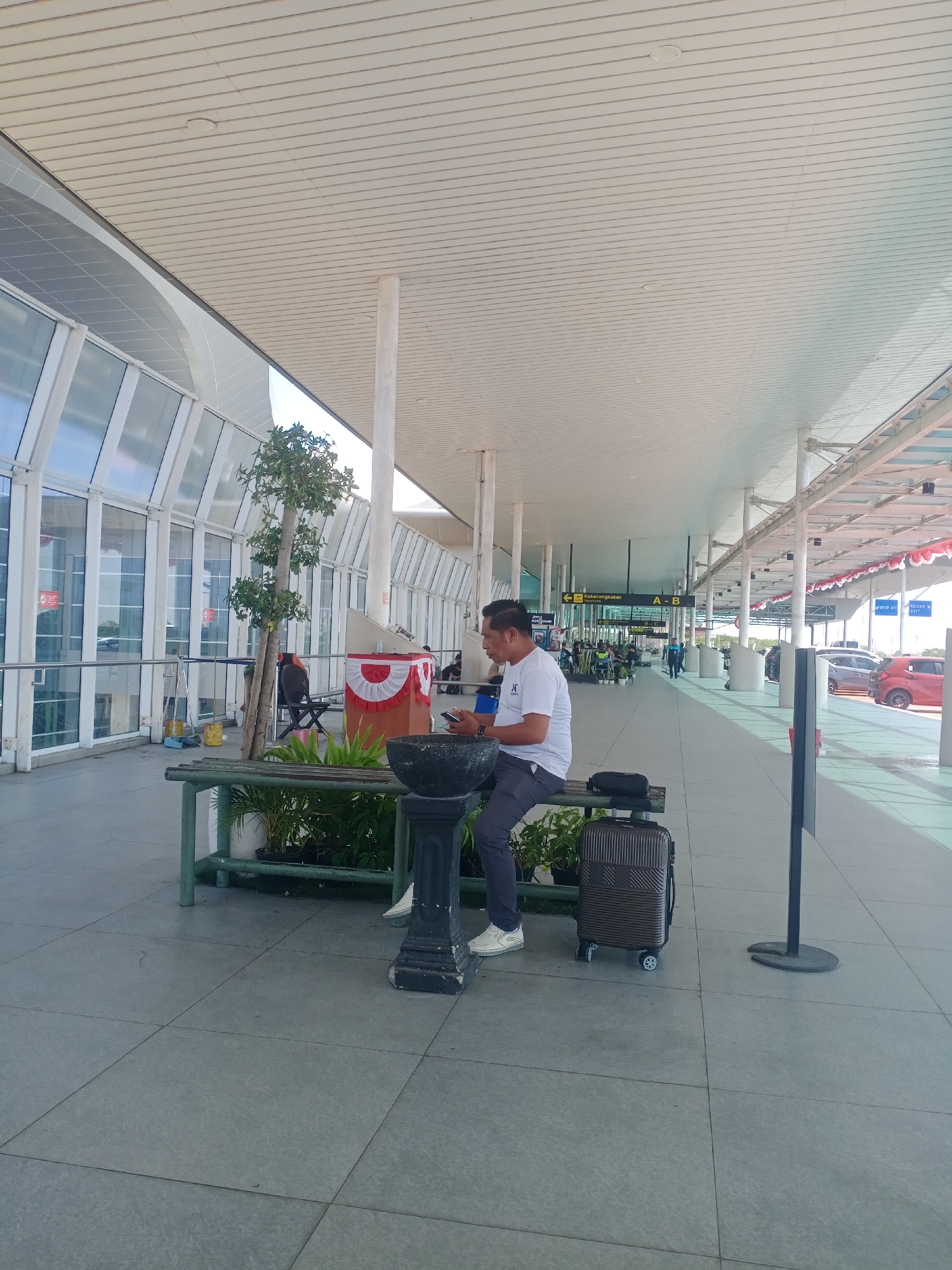 A man in a white t-shirt and grey pants sits on a bench next to a suitcase at what appears to be an airport terminal in Baji Mangngai, Indonesia. He is holding a mobile phone in his hands and looking down at it. The bench is made of wood and metal and is surrounded by potted plants. Behind the bench, there is a large glass window with reflections of cars and trees. The terminal features a white, sloped ceiling with numerous support columns. Signs indicating "Keberangkatan" (Departure) and "A-B" are visible in the distance. Other people and vehicles can be seen in the background. The ground is paved with large grey tiles.