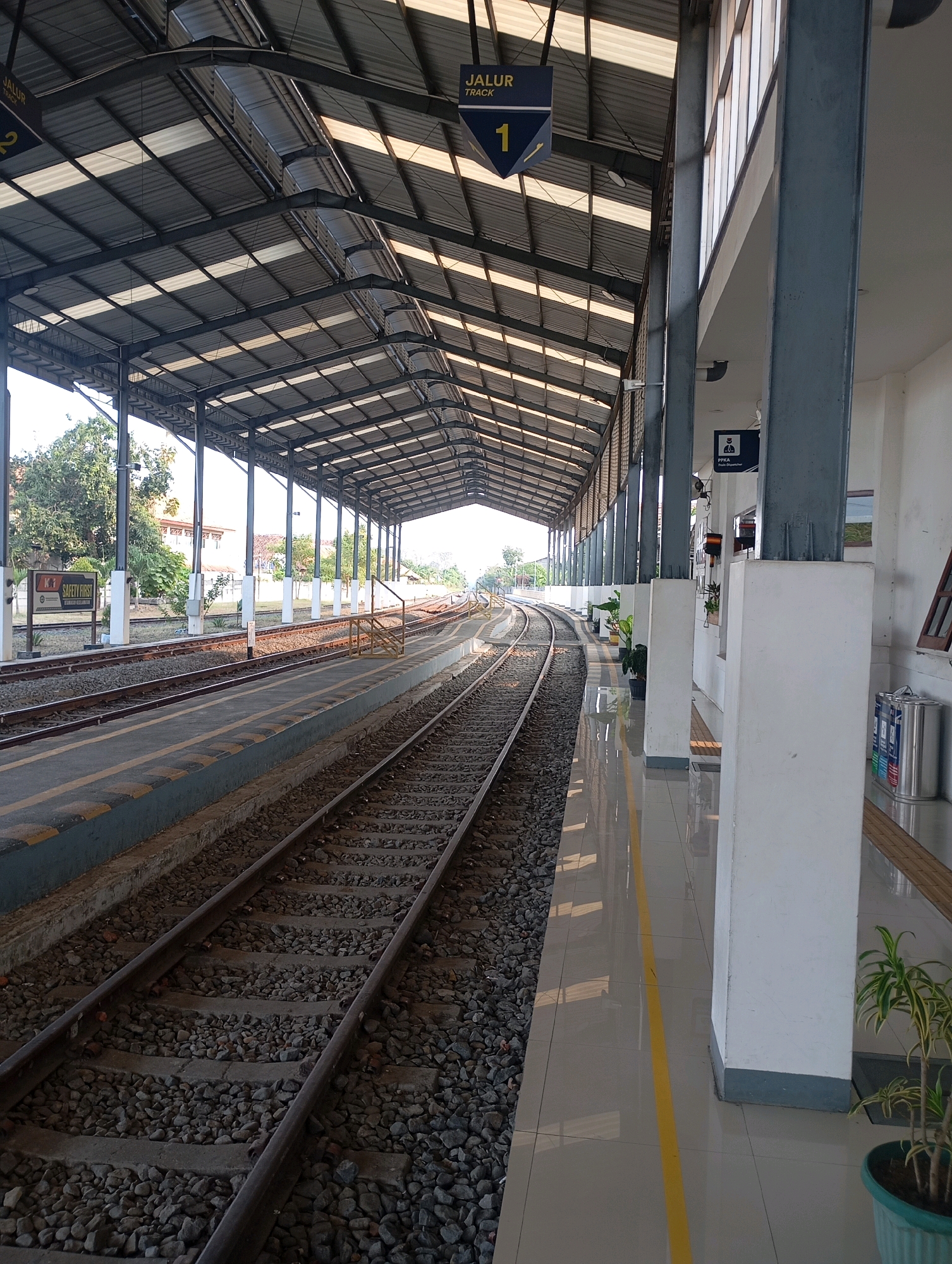 The image shows a train station platform in Slawi, Indonesia. The platform is covered by a metal roof supported by pillars. There are train tracks running alongside the platform. Signs indicate that the track number is 1. A safety first sign is visible on the left. The platform is clean and well-maintained, with yellow lines marking the edge of the platform. There are also some potted plants and trash cans on the platform. The sky is clear and the sun is shining.