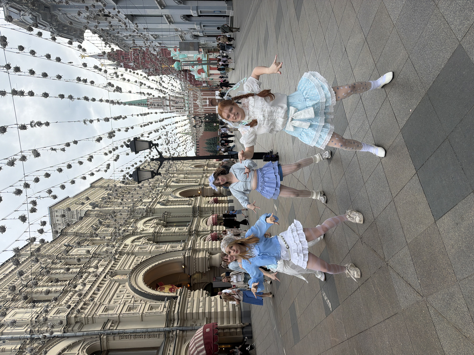 Three young women are standing on a paved street lined with buildings in Moscow, Russia. They are dressed in matching, light blue, Lolita-style dresses with white accents and stockings. Each of them is gesturing with their hands. Behind them is a building with ornate detailing and arched openings, as well as the red walls and towers of the Kremlin. Overhead, strings of decorative lights are strung across the street. Other people are walking in the background.