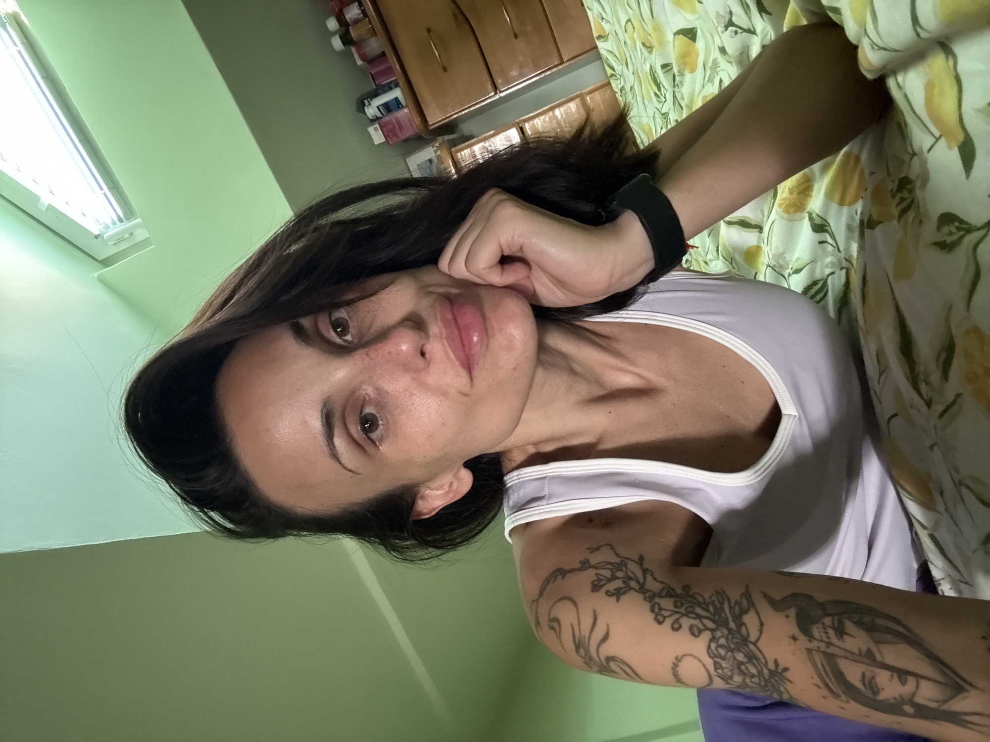 A woman with dark hair, wearing a white tank top and a black fitness tracker on her wrist, is posing for a selfie. Her right hand is near her face, which is slightly tilted. She has a tattoo on her left arm. The background includes a green wall, a window, and a wooden cabinet. A bed with a yellow lemon-patterned bedsheet is also visible. There is no information in the picture to suggest the location is Pleven, Bulgaria.