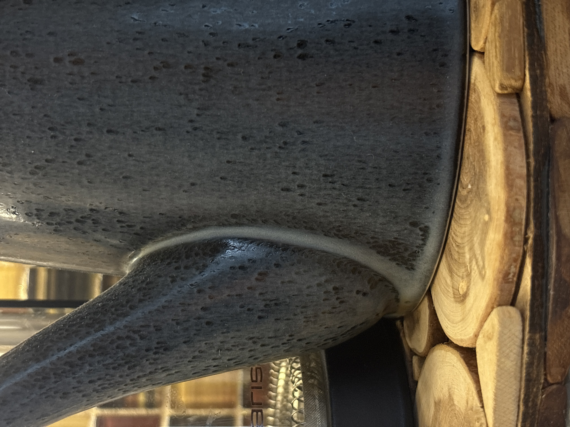 A close-up photograph shows part of a gray, textured object (possibly ceramic or metallic) next to a light brown decorative panel made of small, round wooden slices. The wooden slices are arranged in a stacked, slightly staggered pattern. A blurry reflection is visible in the gray object, showing what appears to be a metal pipe or some sort of fixture. The image likely originated in the city of Chaikovsky, Russia, though this is not directly discernible from the image itself.