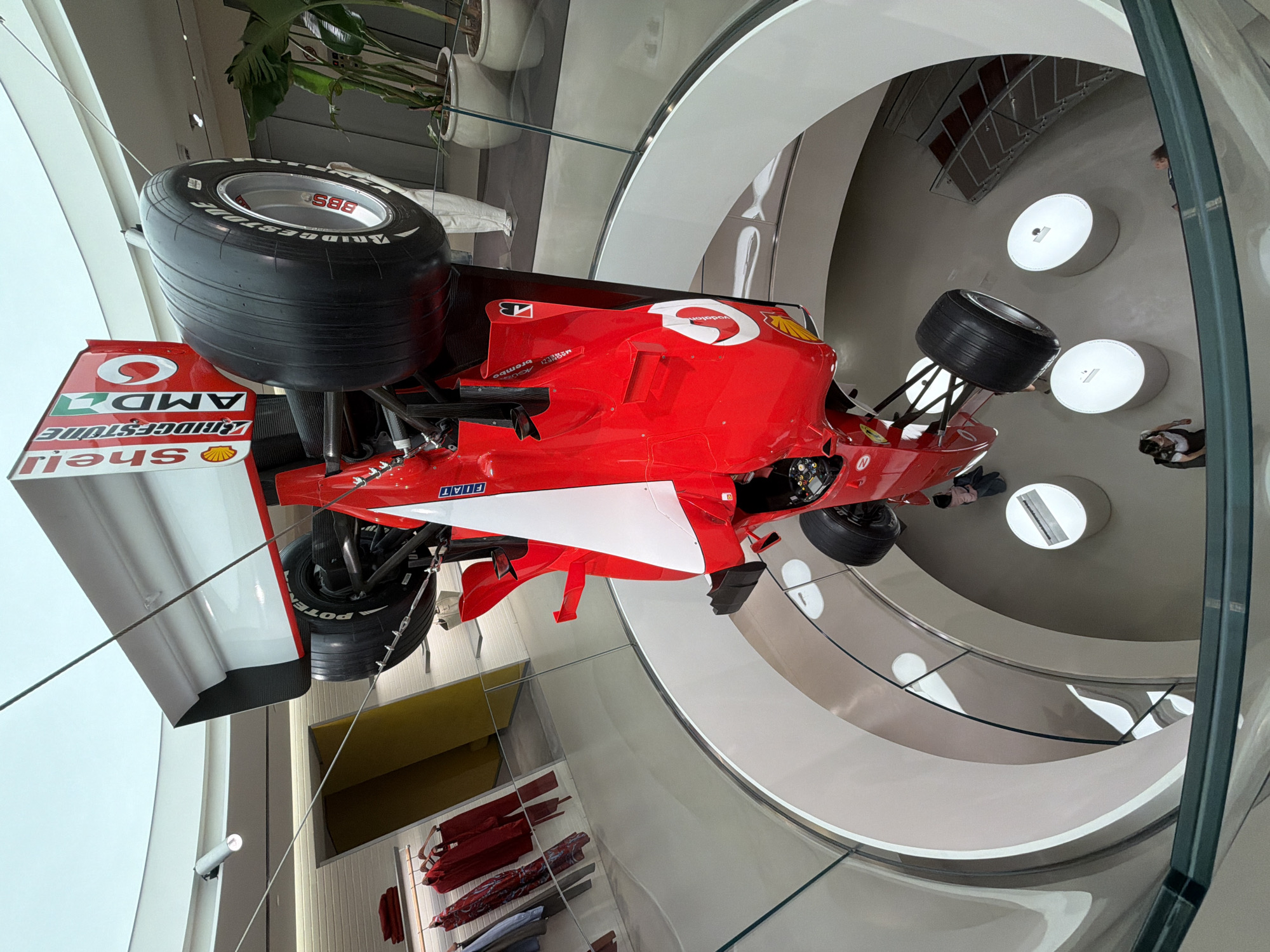 The image depicts a red Ferrari Formula 1 race car on display in a building, presumably a museum. The car features prominent sponsor logos such as Vodafone, Shell, AMD, and Bridgestone. It is suspended in the air, and a glass barrier surrounds it. The background includes a curved, multi-level structure with lighting fixtures. Clothing items on racks are visible on a lower level. A few people can be seen walking in the building. The location appears to be the Ferrari Museum in Milan, Italy.