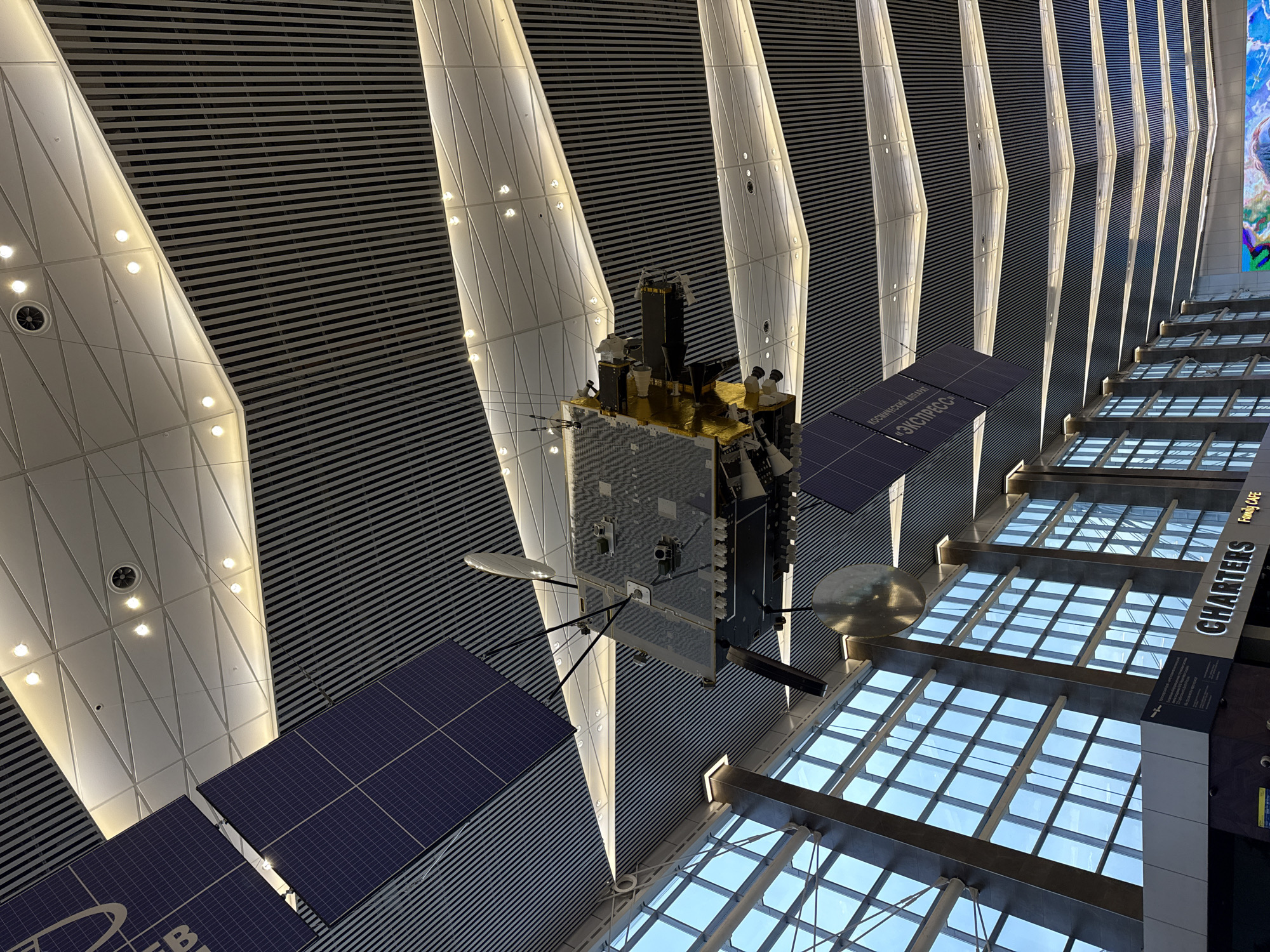 The image shows a low-angle view within the Zhukovsky International Airport (ZIA) near the city of Sukhaya, Russia. An elaborate satellite model hangs from the ceiling, positioned against a backdrop of alternating black and white striped panels interspersed with illuminated, triangular light fixtures.
The satellite model features solar panels and dish-shaped antennas. Adjacent to the model, transparent window structures are visible, allowing daylight to illuminate the interior. Signage for “CHARTERS FAMILY CAFE” is also present, indicating the presence of a food establishment within the airport.