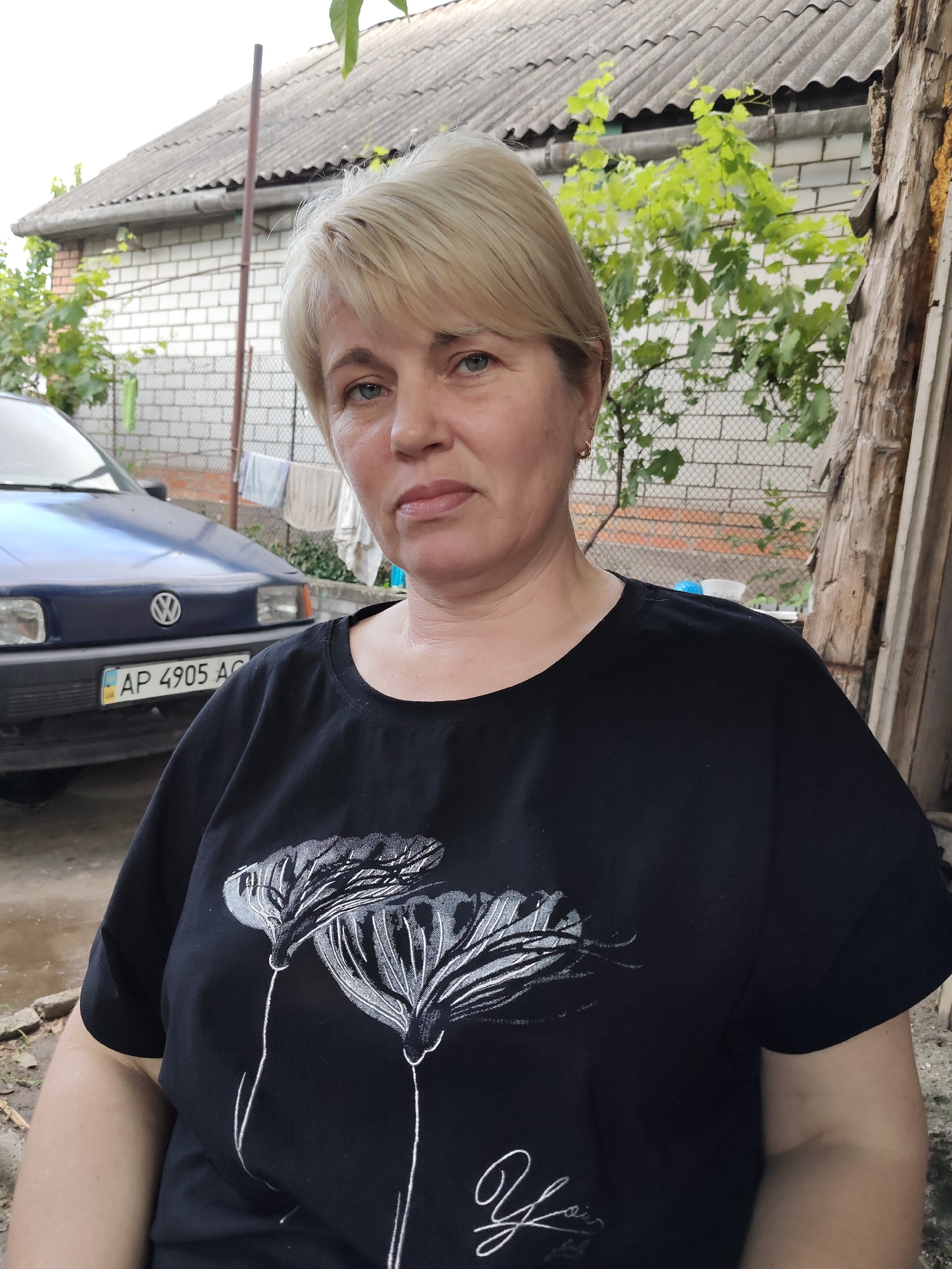 A middle-aged Caucasian woman with blonde hair and blue eyes is pictured outdoors. She is wearing a black t-shirt with a white floral design and is looking directly at the camera. Behind her, a blue car with a Ukrainian license plate (AP 4905 AG) is parked in front of a brick building with a corrugated roof. The environment appears to be a residential backyard, likely in the city of Zaporizhzhia, Ukraine, given the license plate. The overall lighting suggests it is daytime, and the scene has a casual, everyday feel.