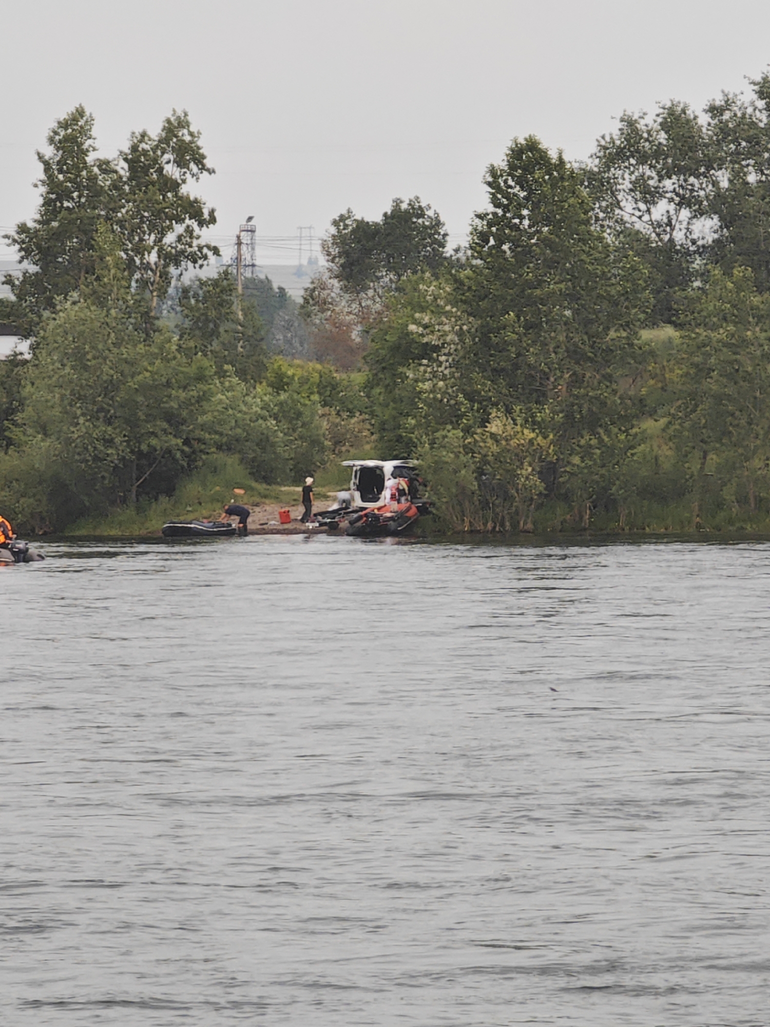 The image depicts a riverbank scene in Peschanka, Russia, with several individuals and watercraft gathered near the shore. The foreground is dominated by a wide body of greyish water with visible ripples. Along the riverbank, which is lined with lush green trees and some sparse vegetation, a white van is parked. Next to the van, on a sandy patch, a group of at least four people are present. One person, wearing a dark shirt, is bending over a deflated inflatable boat. Another person, in a black shirt and white cap, stands near the van. There are two inflatable boats partially visible on the water closer to the shore, one of which appears to have an orange object on it. Further out in the water, another small inflatable boat with at least one person wearing an orange life vest is visible. The background reveals more trees and a hazy sky. In the distance, a tall industrial tower with electrical wires is visible against the overcast sky, suggesting the presence of infrastructure beyond the immediate riverbank. The overall atmosphere suggests an outdoor activity or work being conducted by the river.