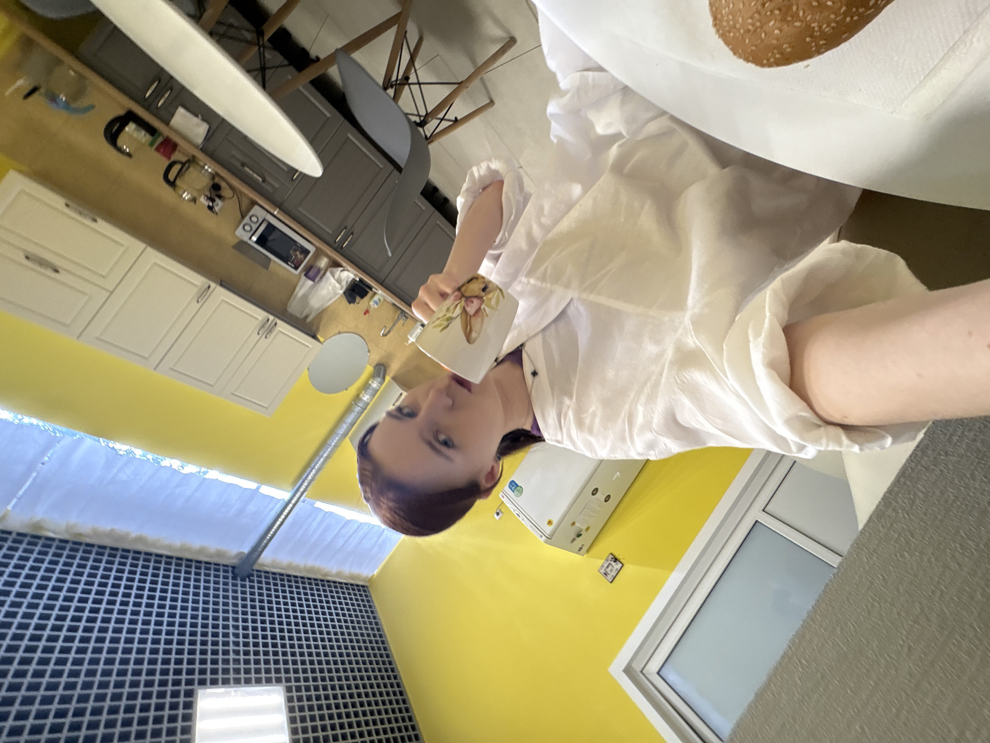 A woman in a white shirt is drinking from a bunny-themed mug in a room with yellow walls, white trim, and a black-and-white tiled wall. A window with white trim is visible. In the background are white cabinets, gray cabinets, a microwave, and hanging utensils. A white ceiling fan and a chair are also visible. The location is Sinyavino, Russia.