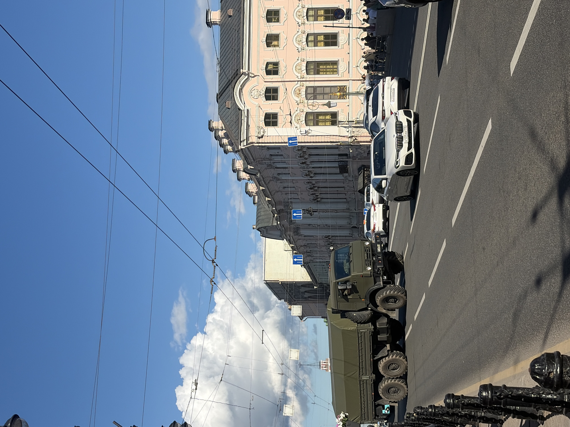 In Saint Petersburg, Russia, a military truck with a green tarp cover is driving on a street. The truck has six wheels and appears to be a heavy-duty vehicle. Several other cars, including white sedans, are also on the road. In the background, a long, pink building with ornate architecture and arched windows is visible. There are pedestrians on the sidewalk in front of the building. The sky is blue with some white clouds, and power lines are strung across the scene. The image captures a typical urban street scene with a mix of civilian and military vehicles.