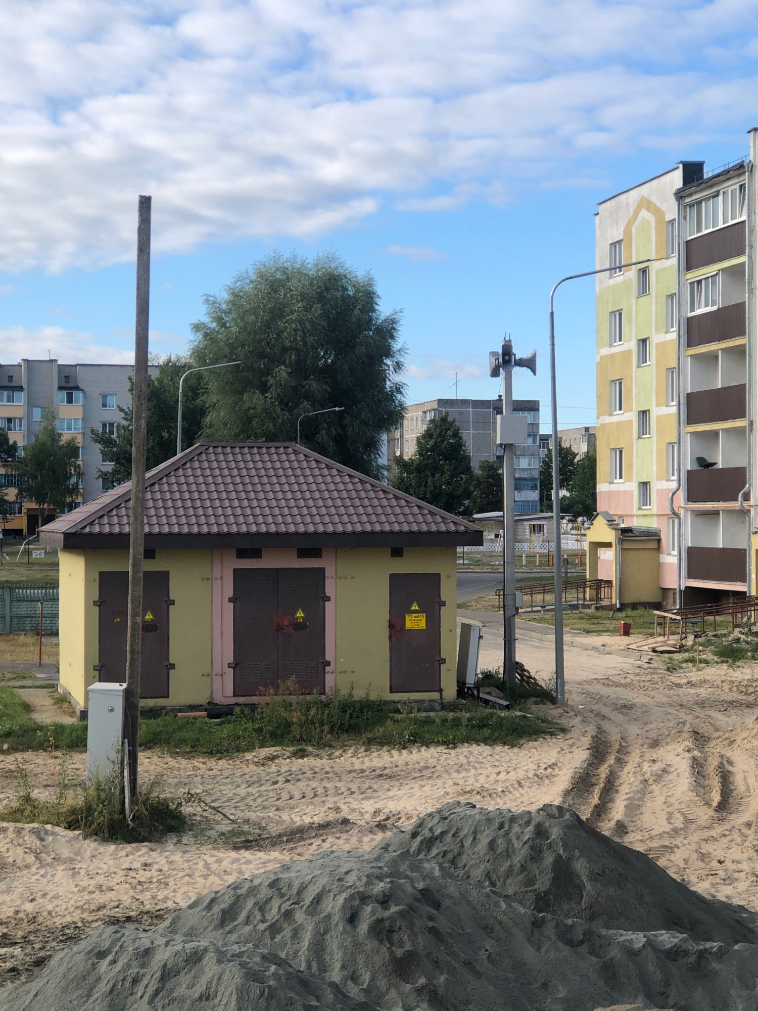 The image shows a small, yellow transformer substation building with a brown, tiled roof in Zhytkavichy, Belarus. Two large, brown doors with yellow high-voltage warning signs are visible on the front of the building. A tall, gray utility pole with what appears to be a siren or loudspeaker mounted on it stands to the right of the substation. In the background, there are several multi-story apartment buildings, some with colorful facades. The ground around the substation is covered with sand and dirt, with tire tracks indicating recent vehicle activity. A large pile of gray gravel or sand is in the foreground, suggesting ongoing construction or landscaping. Lush green trees are scattered throughout the scene, and the sky is partly cloudy with a bright blue sky. No people are visible in the image.