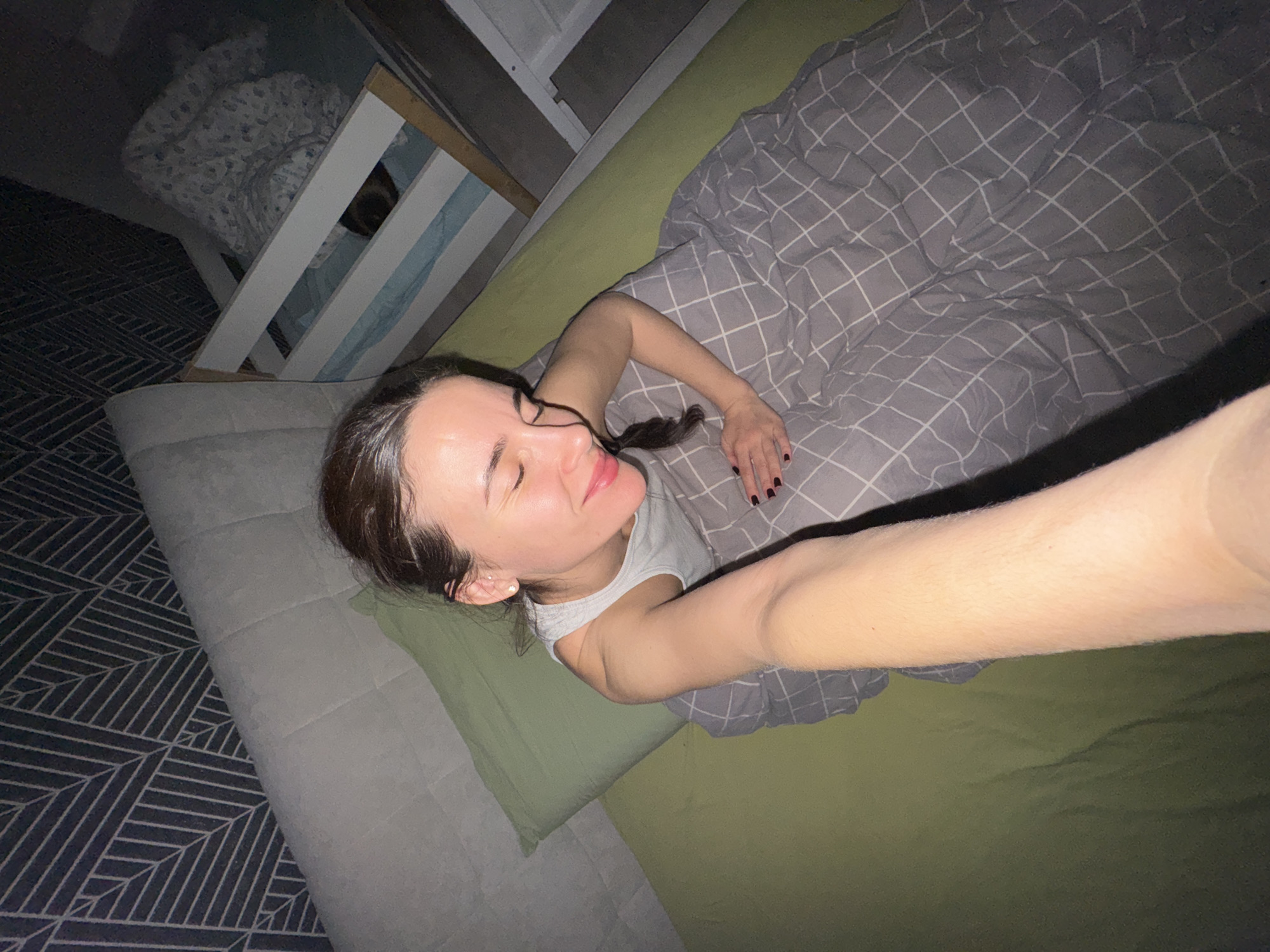 A woman lies on a bed, covered partially by a gray patterned blanket. She rests on a green pillow. A gray couch sits adjacent to the bed. The room appears dimly lit. A child's crib is partially visible in the background. The woman is taking a selfie. The location is indicated to be in the city of Chaikovsky, Russia.
