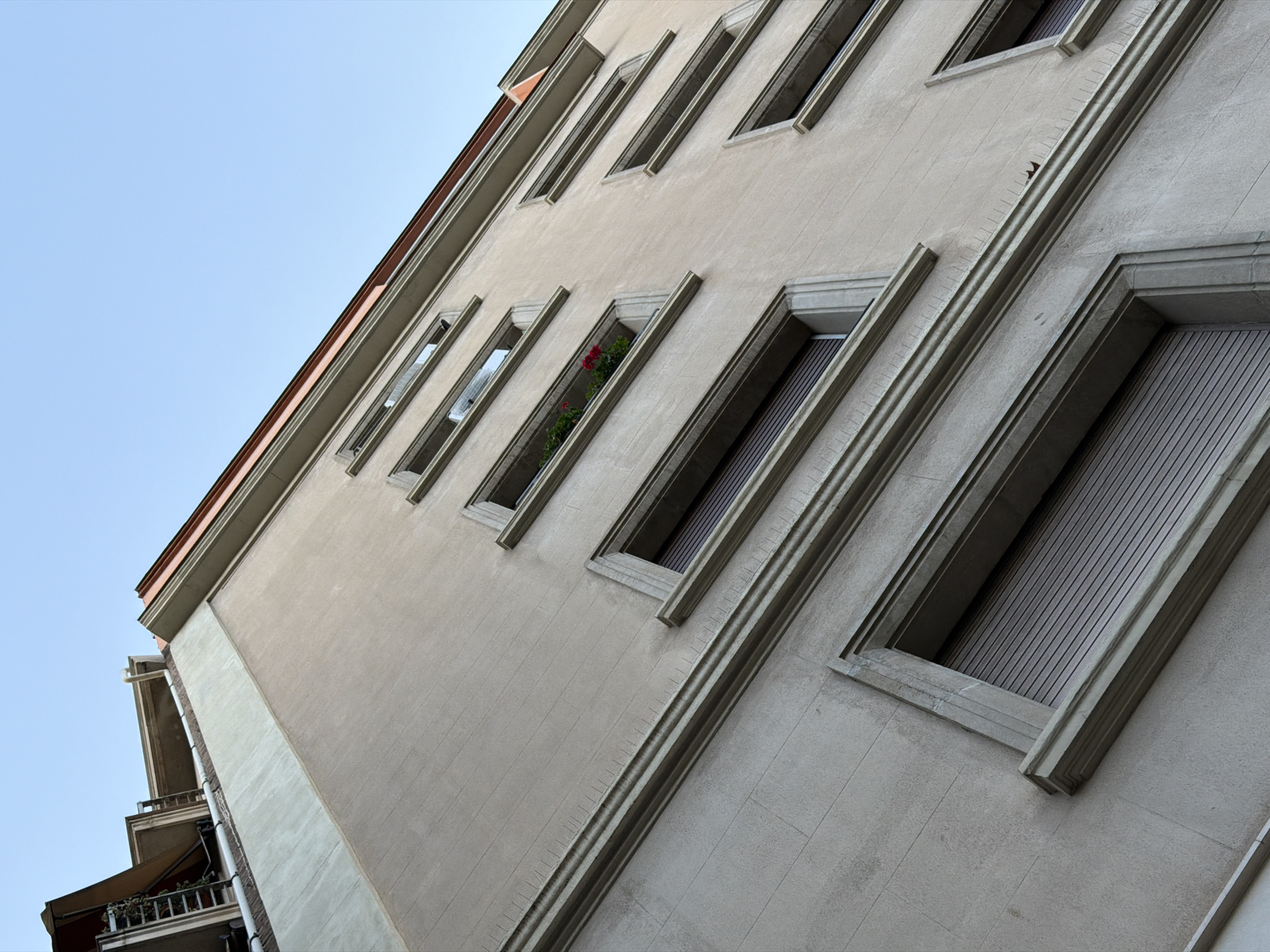 This is a low-angle, wide shot of the exterior of a light-colored building in Barcelona, Spain. The building has a stucco finish and appears to be multi-story, with several windows visible. The windows are rectangular and recessed into the facade, framed by decorative molding. Some windows have exterior shutters that are closed, revealing horizontal slat patterns. One window on a lower floor contains a planter with red flowers. The roofline is visible at the top of the image, with a terracotta-colored coping and a light-colored overhang. The sky above is clear and light blue. The architecture suggests a residential or mixed-use building in a European city. The image is an architectural detail shot, focusing on the facade and its features. There are no people or vehicles visible in the image. The overall impression is one of urban architecture and a clear day.