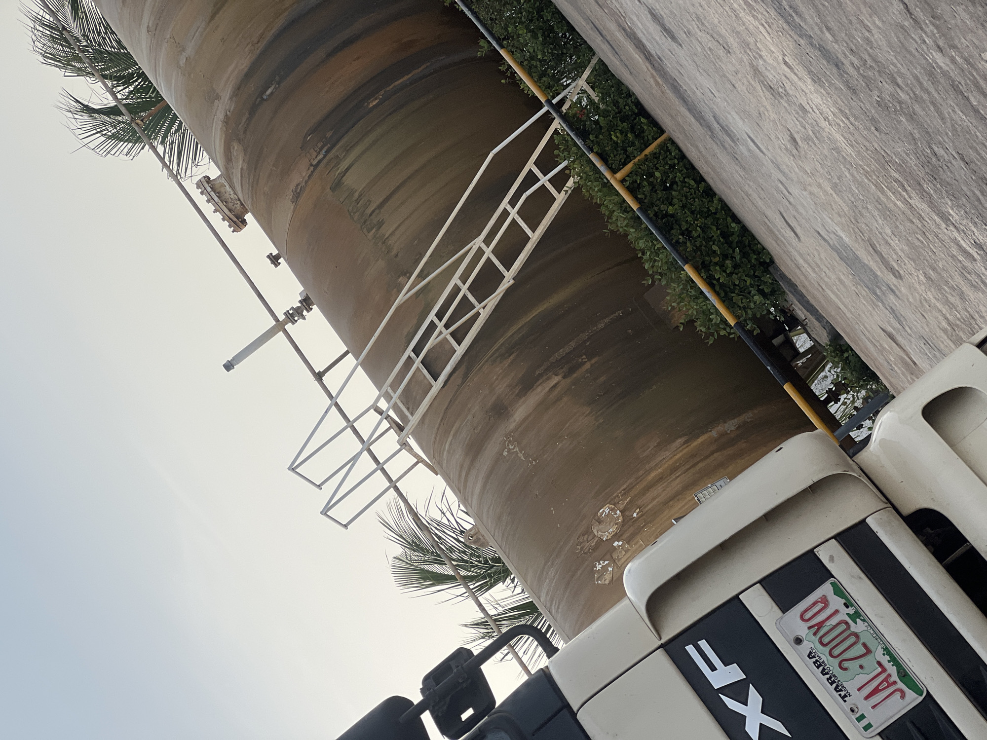The image shows a close-up of the side of a large, cylindrical tank, likely a tanker truck. The tank is a dull brown color and appears to be made of metal. A white metal ladder is attached to the side of the tank, extending upwards. In the foreground, the front of a white truck is visible, with a license plate that reads "JAL-200YO TARABA" and "NATURE'S GIFT TO THE NATION". The truck has the letters "XF" prominently displayed. The scene is set outdoors, with some green foliage visible behind the tank. The sky is a pale, overcast grey. The image is oriented such that the truck and tank are at an angle, suggesting the photo was taken from a low vantage point or the camera was tilted. There are no people visible in the image. The overall impression is of industrial equipment in an outdoor setting, possibly a depot or storage area.