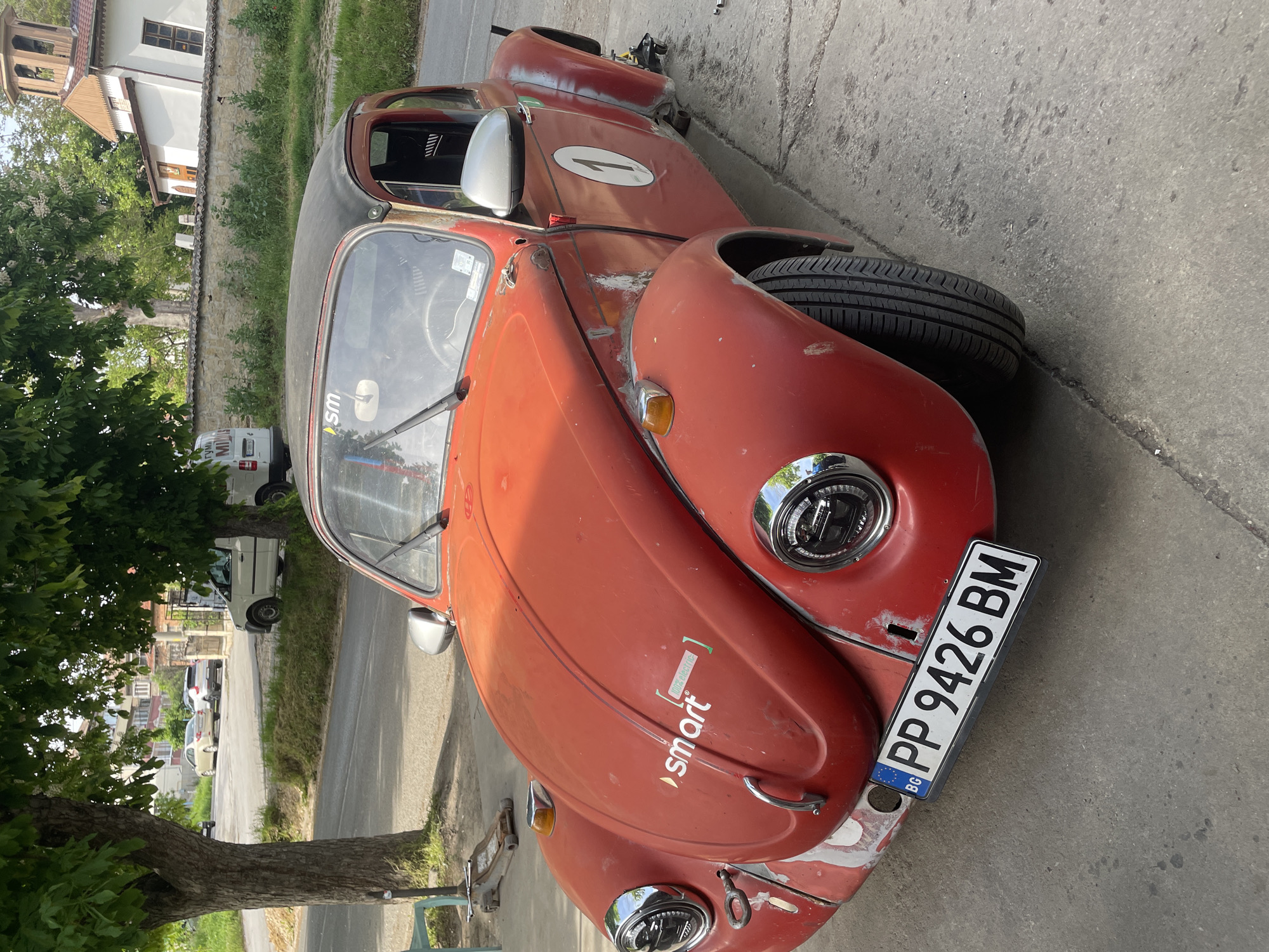 A red, heavily modified and partially restored classic Volkswagen Beetle is parked on a grey concrete sidewalk in a residential area of Razgrad, Bulgaria. The car has a number "1" painted on its passenger side, a "smart" sticker on the hood, and a license plate reading "PP 9426 BM" from Bulgaria. The car's front end shows signs of repair work, with exposed areas. A tree is present on the left side of the image. In the background, there are residential buildings and other vehicles parked along the street.