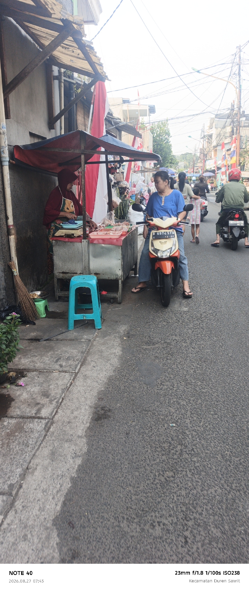 In East Jakarta, Indonesia, a street scene unfolds with a food vendor and passing traffic. A woman in a maroon hijab and apron stands behind a cart laden with raw chicken pieces, some arranged in pink plastic trays. She appears to be preparing or selling the meat. In front of the cart, a bright blue plastic stool sits on the concrete sidewalk. To the left, a broom leans against the wall of a building.
A woman wearing a blue t-shirt and jeans is seated on an orange and white scooter, facing towards the viewer and the food stall. Her posture suggests she might be waiting or has stopped to observe the vendor. Further down the street, several other people are visible on scooters, with one rider in a green jacket and red helmet heading away from the camera. The street is paved with asphalt, and the background shows buildings and utility poles with numerous wires. The day appears to be bright, casting shadows.