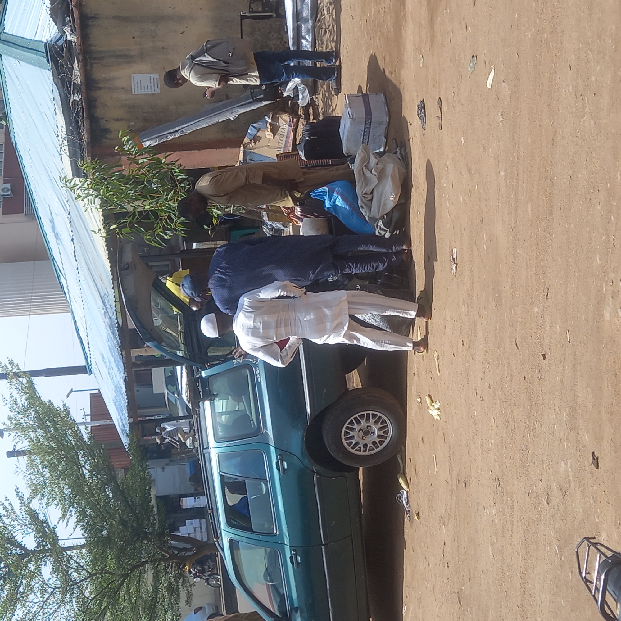 A partly obscured green station wagon is parked on a dirt surface next to a building with a light blue corrugated metal roof. Four people are visible near the vehicle, seemingly loading items onto it. One man wears a white outfit and head covering, while another is dressed in dark blue. The other two people are further away and obscured by the people in front. Assorted luggage, boxes, and bags are on the ground near the car. A motorcycle or scooter is partially visible near the lower left corner. The location is Dadi Riba, Nigeria.