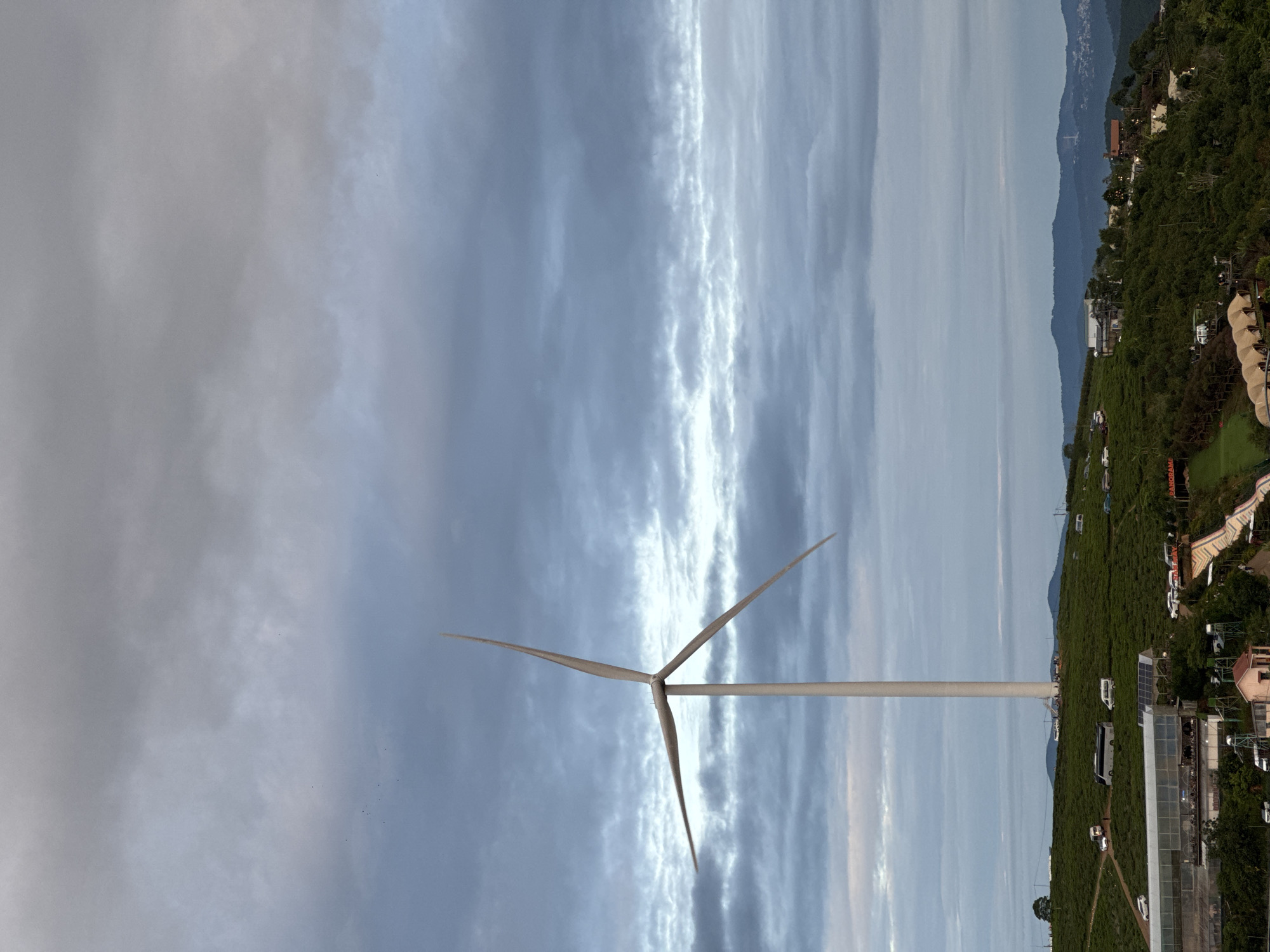 A tall wind turbine stands prominently against a cloudy sky. The blades of the turbine are positioned as if to cut into the upper portion of the frame. The turbine is set against a backdrop of a landscape that includes trees, some buildings, and distant mountains. This appears to be in or around the city of Phát Chi, Vietnam, given the nature of the terrain and architecture, though no explicit identifiers are visible. The image is taken from a low angle, making the turbine appear larger. The ground near the base of the turbine seems to contain a mix of vegetation and buildings.