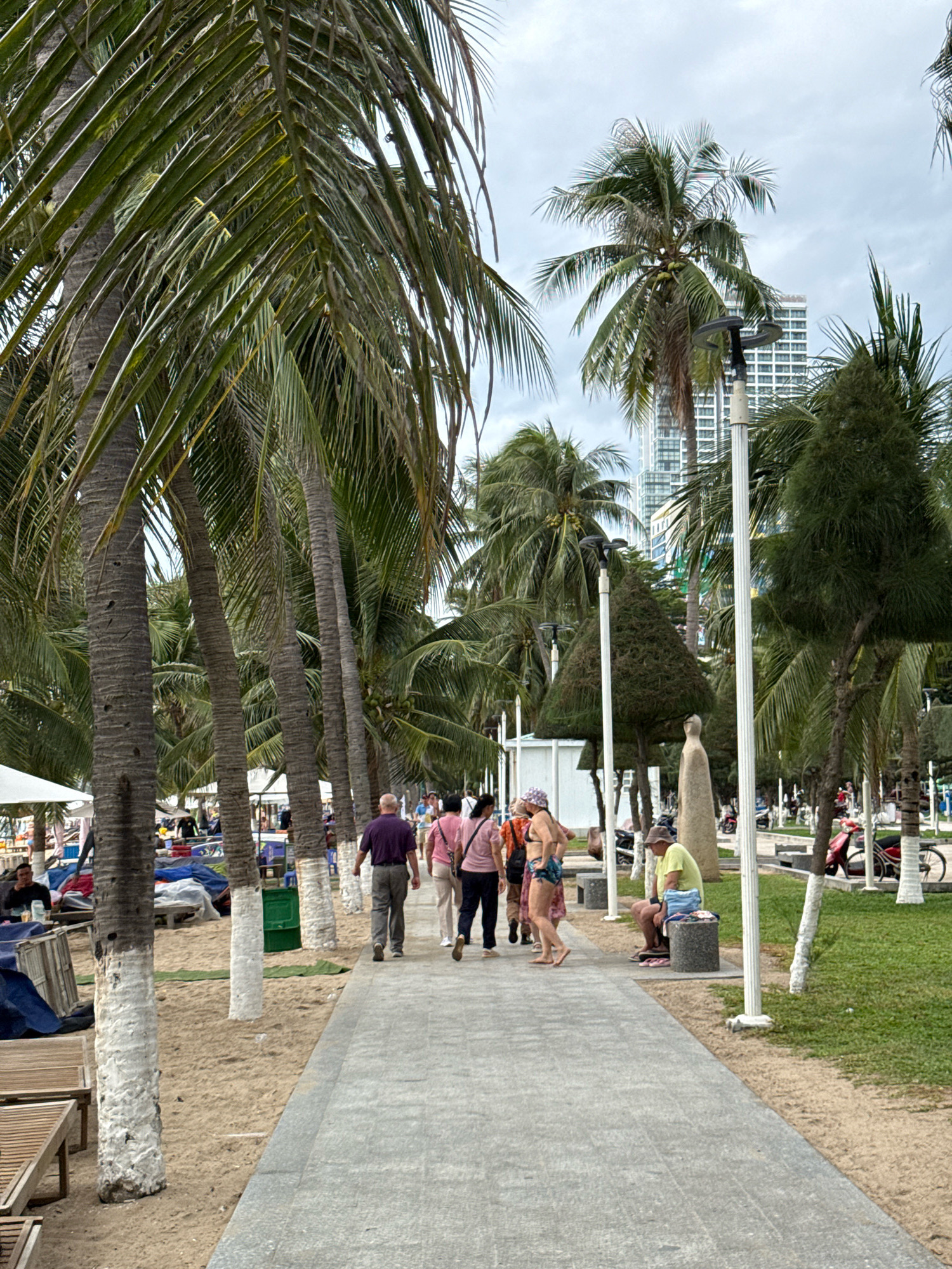 This image depicts a sidewalk lined with palm trees along a beach in Nha Trang, Vietnam. In the foreground, a gray concrete sidewalk runs diagonally from the bottom right to the top left of the frame. To the left of the sidewalk is a sandy beach with several lounge chairs. Further back, on the right side of the sidewalk, there is a grassy area with more trees and people. In the background, there are tall buildings, indicative of a city. Several people are walking along the sidewalk, some are sitting on benches, and others are visible on the beach in the distance. The sky is overcast.