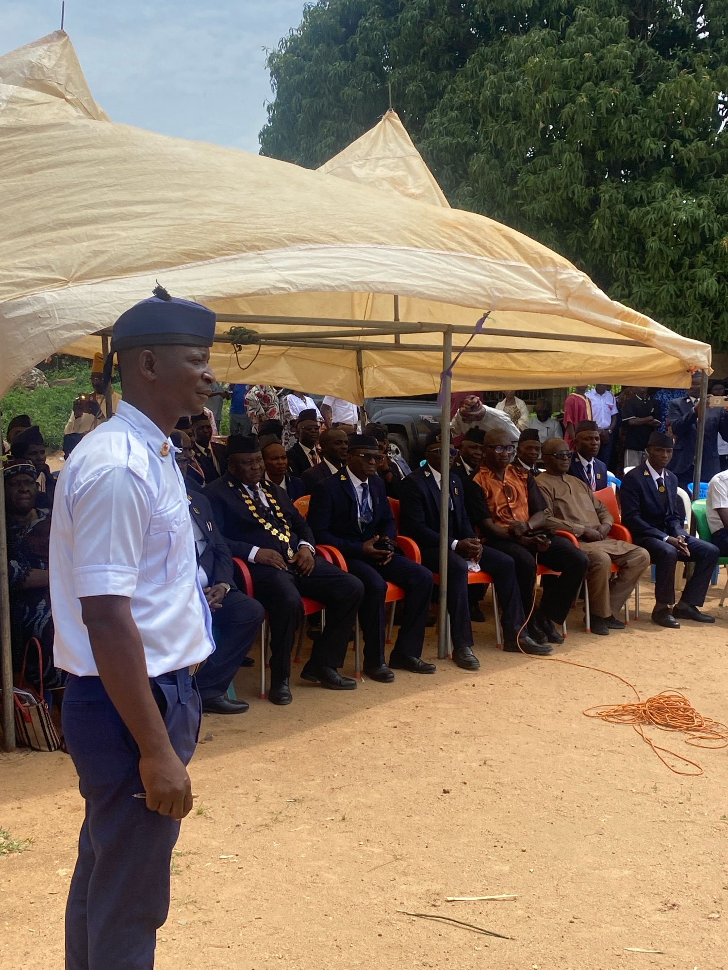 In Binchi, Nigeria, this outdoor scene captures a gathering under a large, beige tent. In the foreground, a man in a white uniform shirt and dark blue pants, wearing a dark blue peaked cap with a yellow emblem, stands to the left, facing right and looking away. His left hand is at his side, holding a pen. Behind him, several men are seated in chairs, some wearing dark suits and ties, others in traditional African attire. One man wears a ceremonial gold chain. The ground is covered with reddish-brown dirt. To the right of the seated men, a coil of orange electrical cord lies on the ground. Lush green trees form a backdrop behind the tent. The sky is overcast.