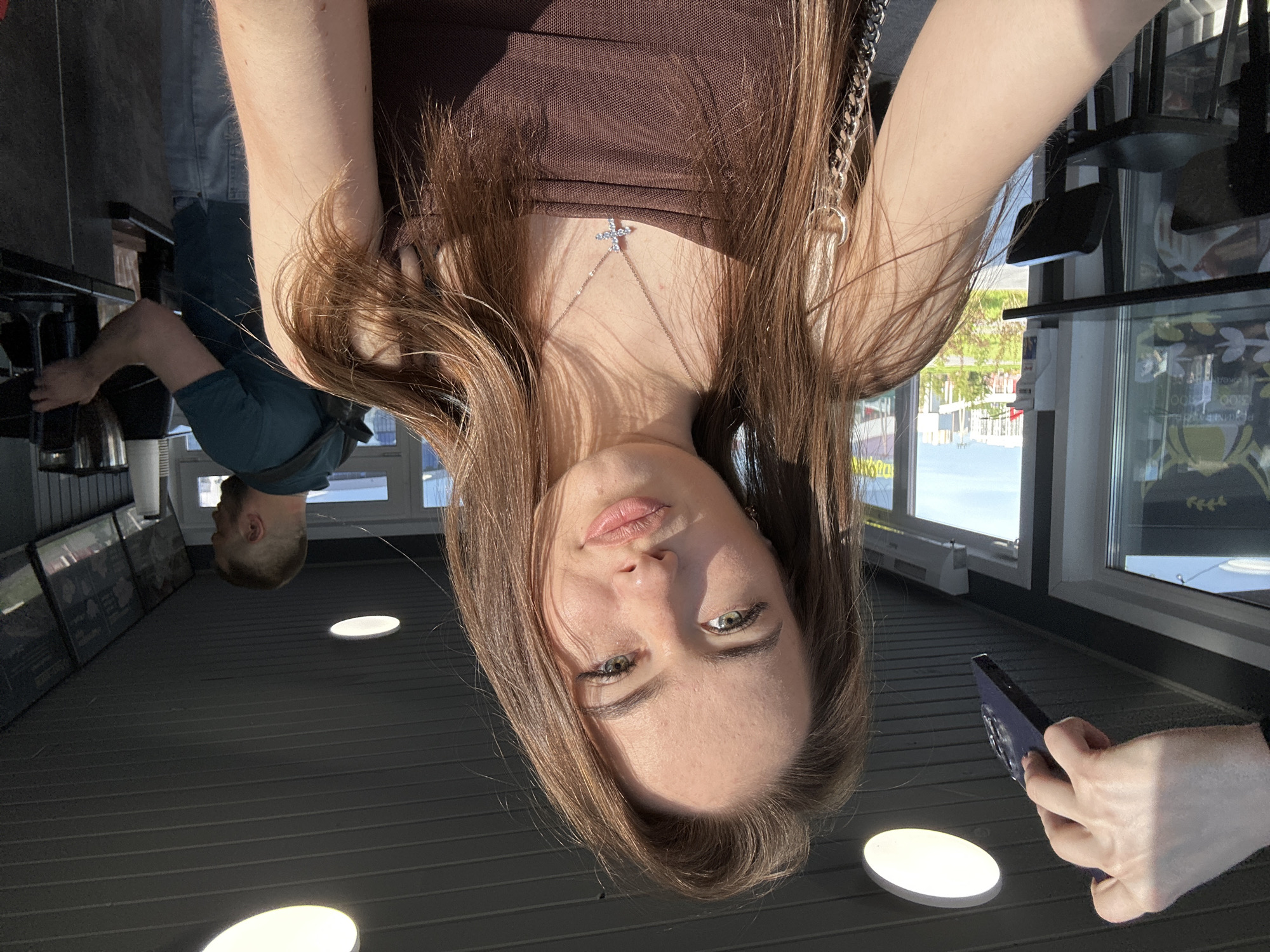 The image depicts an indoor scene, likely a cafe or similar establishment, brightly lit by natural sunlight streaming through large windows. The primary subject is a young woman, captured in what appears to be a selfie, as she is holding a dark-colored smartphone in her right hand. She has long, straight brown hair, light skin, and striking green eyes, with her lips slightly parted. She wears a brown top and a silver chain necklace featuring a small, glittering cross pendant at the front.
In the background, another person, a man with short, light hair, is visible, possibly working behind a counter or bar area. He is wearing a dark blue or teal shirt. The interior features a dark-colored ceiling with horizontal paneling and several bright, recessed, oval-shaped lights. On a wall to the left, there are several framed items, possibly posters or artwork.
Through the large windows, an outdoor scene is visible, suggesting it is daytime and sunny. There are hints of greenery outside, and possibly other structures or roads. Some indistinct text and graphics are faintly visible on the windowpane, including what might be "13:00" and "00" in one section. The woman's left arm also features a silver chain accessory. The overall mood is casual and bright.
