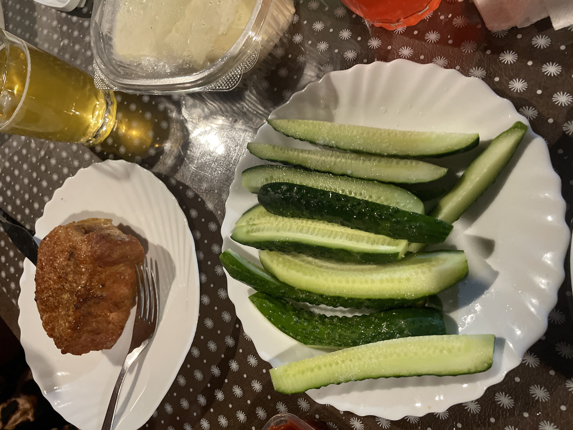 The image shows a close-up of a table set for a meal. In the foreground, a white, scalloped plate is filled with sliced cucumbers, some of which are glistening with water droplets. To the left of the cucumbers, a small red object, possibly ketchup, is partially visible in a small dish. In the background, a round white plate holds a golden-brown, fried cutlet or meatball, with a fork resting on the plate. Next to this, a glass of amber-colored liquid, likely beer, sits on the table, casting a reflection. A clear plastic container filled with a light-colored substance, possibly mashed potatoes or dip, is also visible. The table surface is covered with a brown tablecloth featuring a white starburst pattern. The lighting appears to be artificial, casting shadows on the table. The scene suggests a casual meal, likely at home.