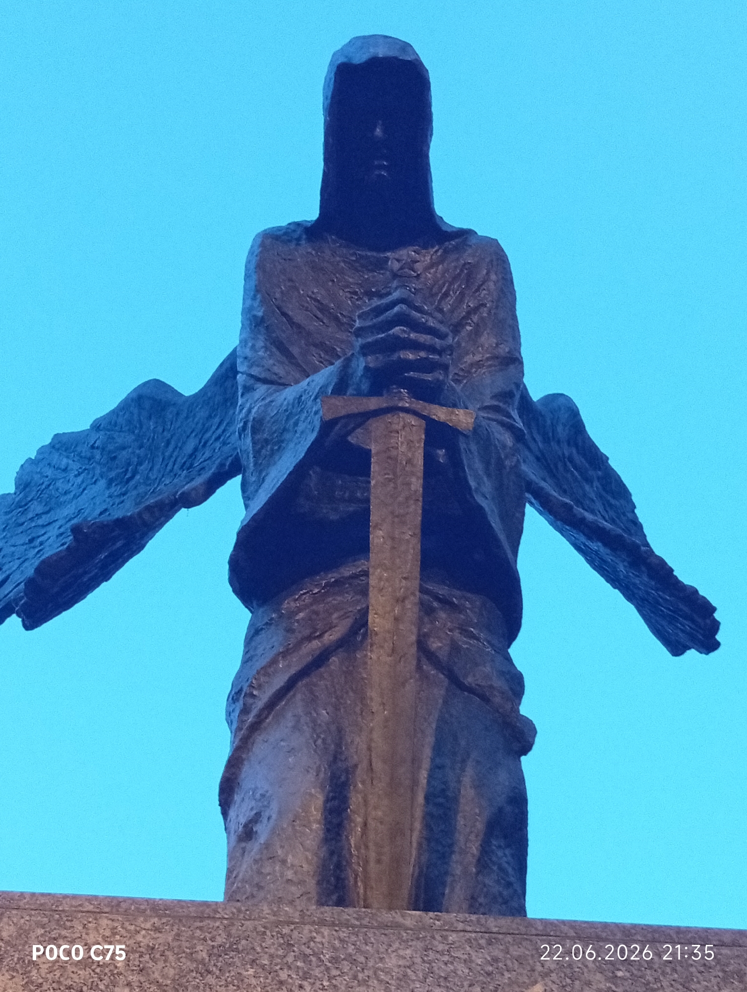 The image shows a close-up, low-angle shot of a dark bronze statue of an angel with wings spread wide. The angel is depicted with its hands clasped around a sword that runs vertically in front of its body. The figure wears a hooded robe that obscures most of its facial features, giving it a solemn and imposing appearance. The statue stands on a granite pedestal. The background is a clear, bright blue sky, suggesting the photo was taken during the day. In the bottom left corner, "POCO C75" is visible, indicating the phone model used. In the bottom right corner, a timestamp reads "22.06.2026 21:35," which is a future date and likely a photo editing error or a placeholder. This statue is the Archangel Michael located in Wroclaw, Poland, specifically at the Archangel Michael's Chapel in the district of Nowy Dwór.